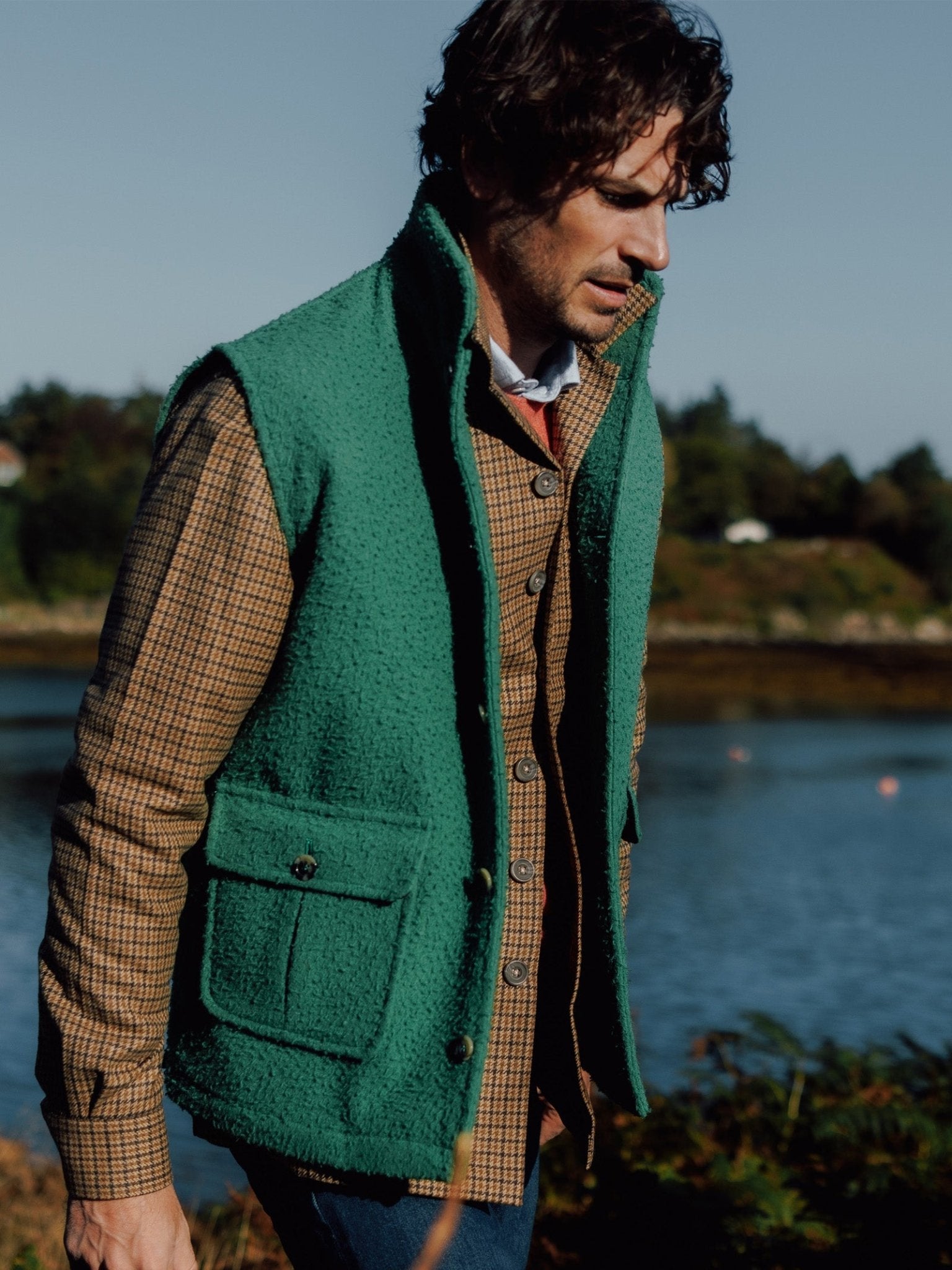 A man with wavy dark hair wears a green Campbells of Beauly Casetino Fleece Gilet over a brown plaid shirt, standing outdoors by the water with trees and houses in the background on a sunny day.