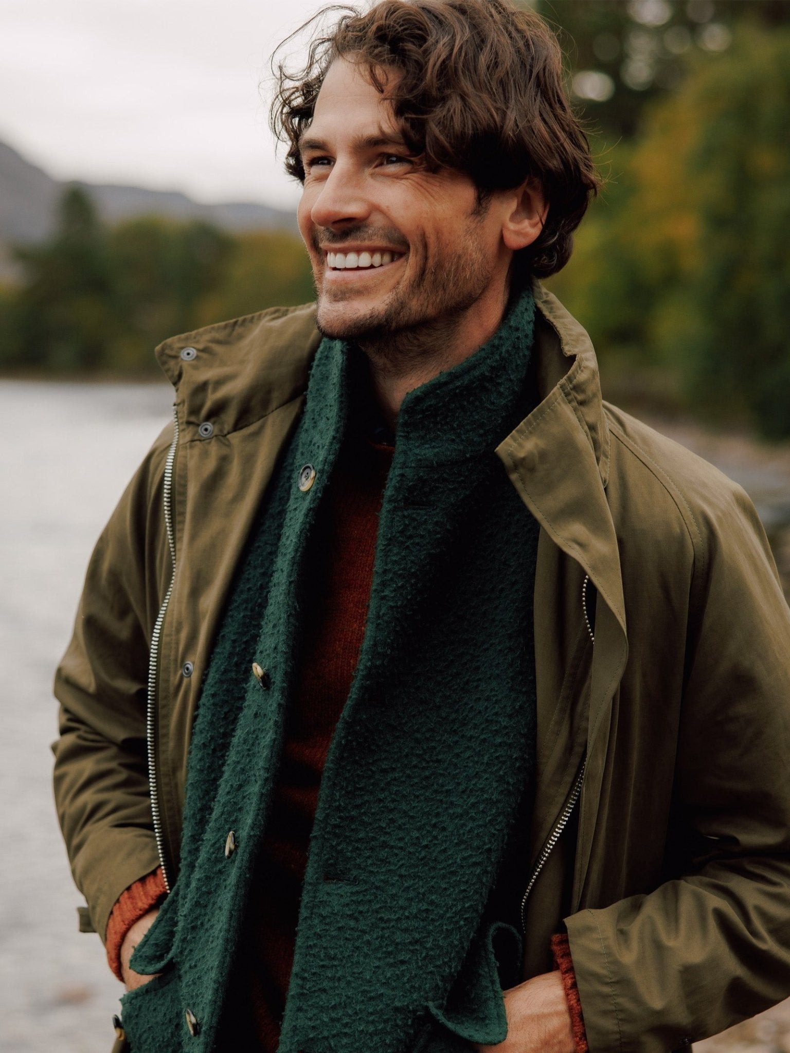 A man with wavy brown hair smiles by the water, wearing a Campbells of Beauly Casetino Fleece Gilet in olive green, paired with a dark green scarf and rust sweater. Trees and distant mountains blur in the background.