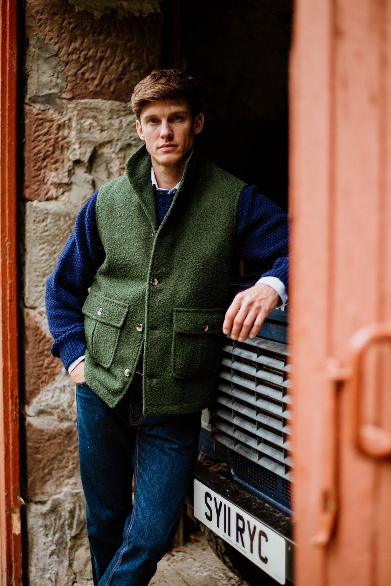 A man wearing a green Campbells of Beauly Casetino Fleece Gilet and blue sweater leans against a parked vehicle with a visible license plate, standing in a doorway framed by stone and wood.