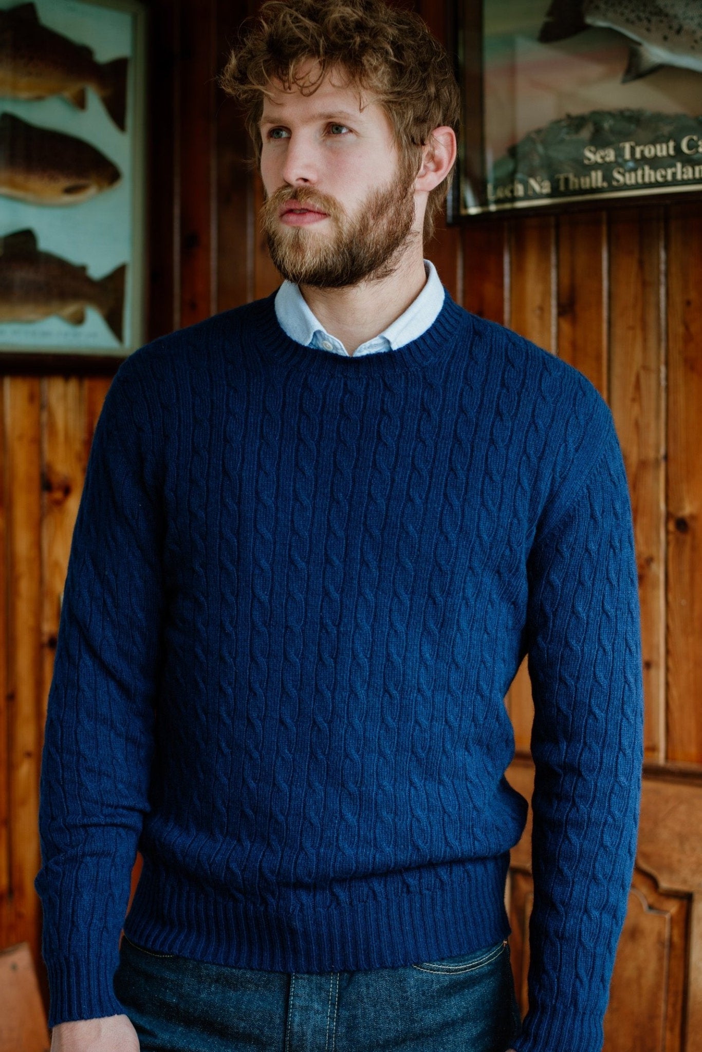 A man with curly hair and a beard stands indoors wearing the Campbells of Beauly Cashmere Cable Crew Jumper over a collared shirt, set against wooden paneling and framed fish prints that evoke the rustic charm of the Scottish Borders.