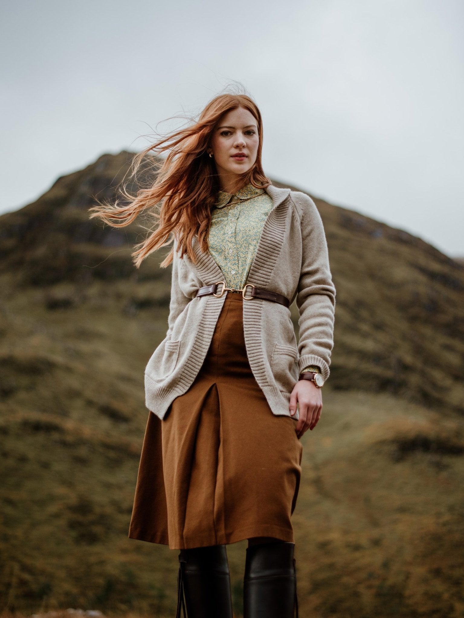 A woman with long red hair stands outdoors before a grassy hill, her hair blown by the wind. She wears a Campbells of Beauly Cashmere Shawl Cardigan, a green patterned blouse, brown skirt, and black boots.