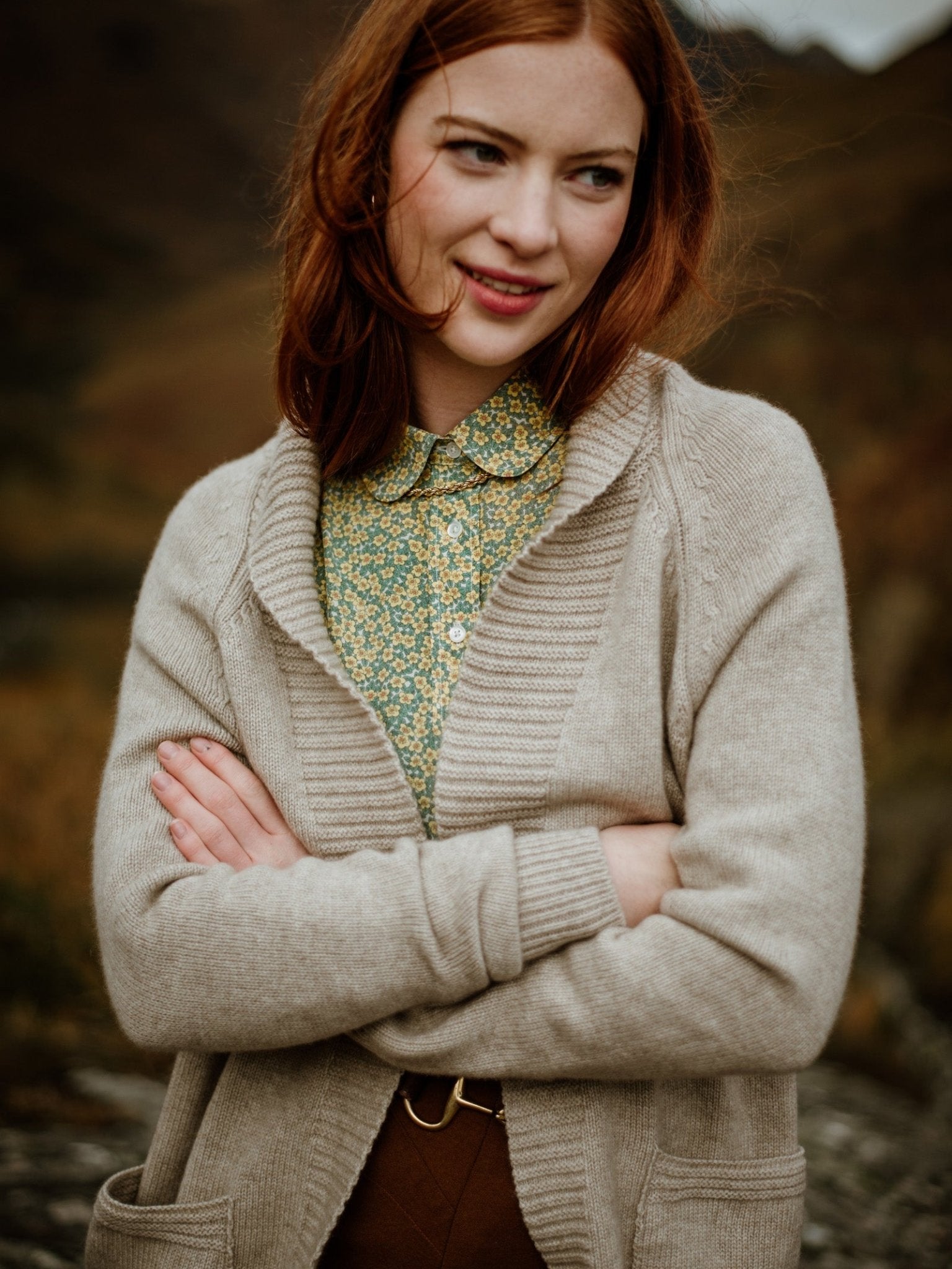 A young woman with fair skin and long red hair stands outdoors, wearing the Campbells of Beauly Cashmere Shawl Cardigan over a green patterned blouse. Smiling softly, she crosses her arms as the nature-filled background blurs behind her.