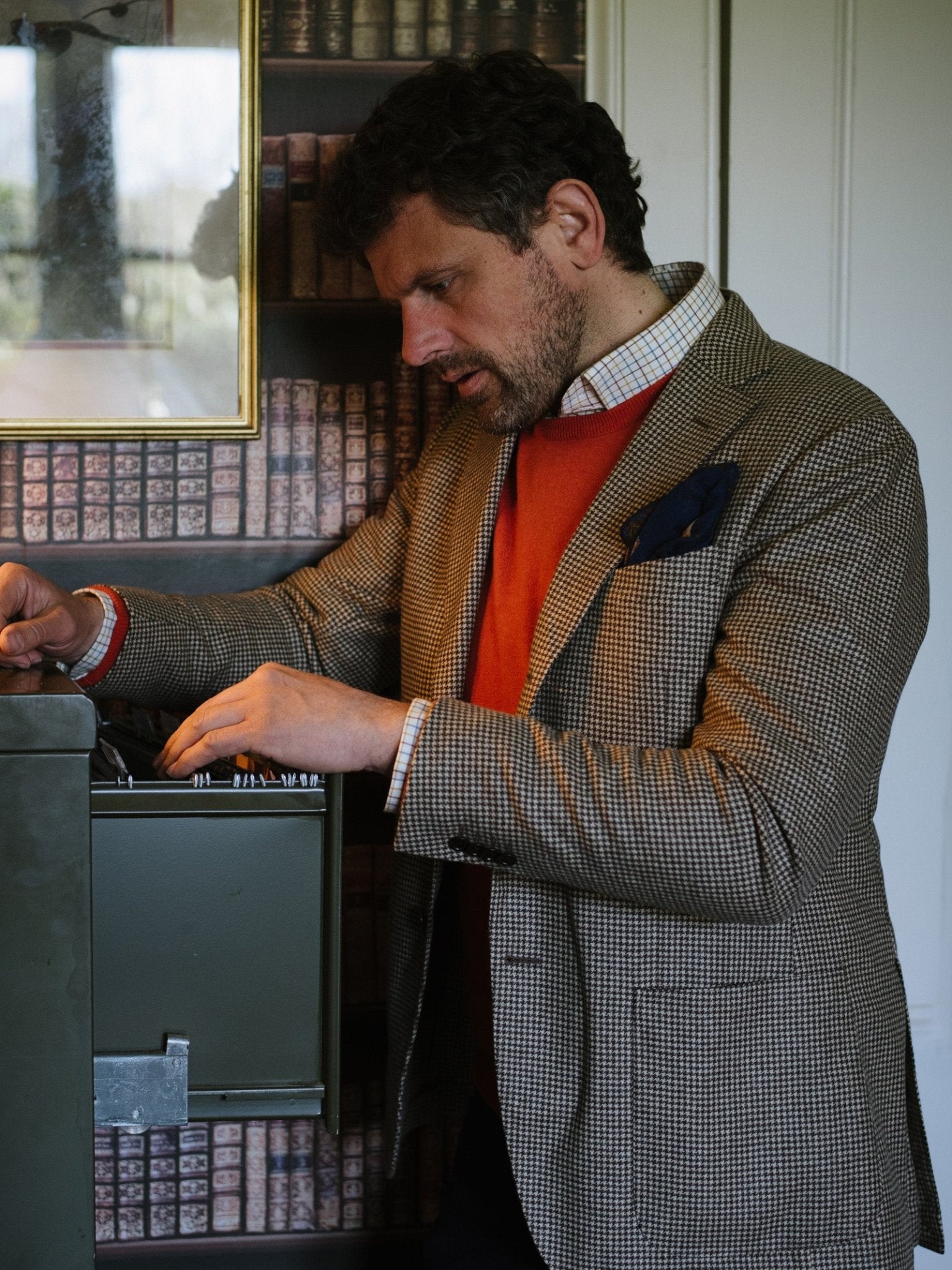 A man wearing a Campbells of Beauly Cashmere Crew Neck Jumper and a checked blazer stands indoors, intently examining a metal box with a luxe handle. Bookshelves filled with books appear in the background.