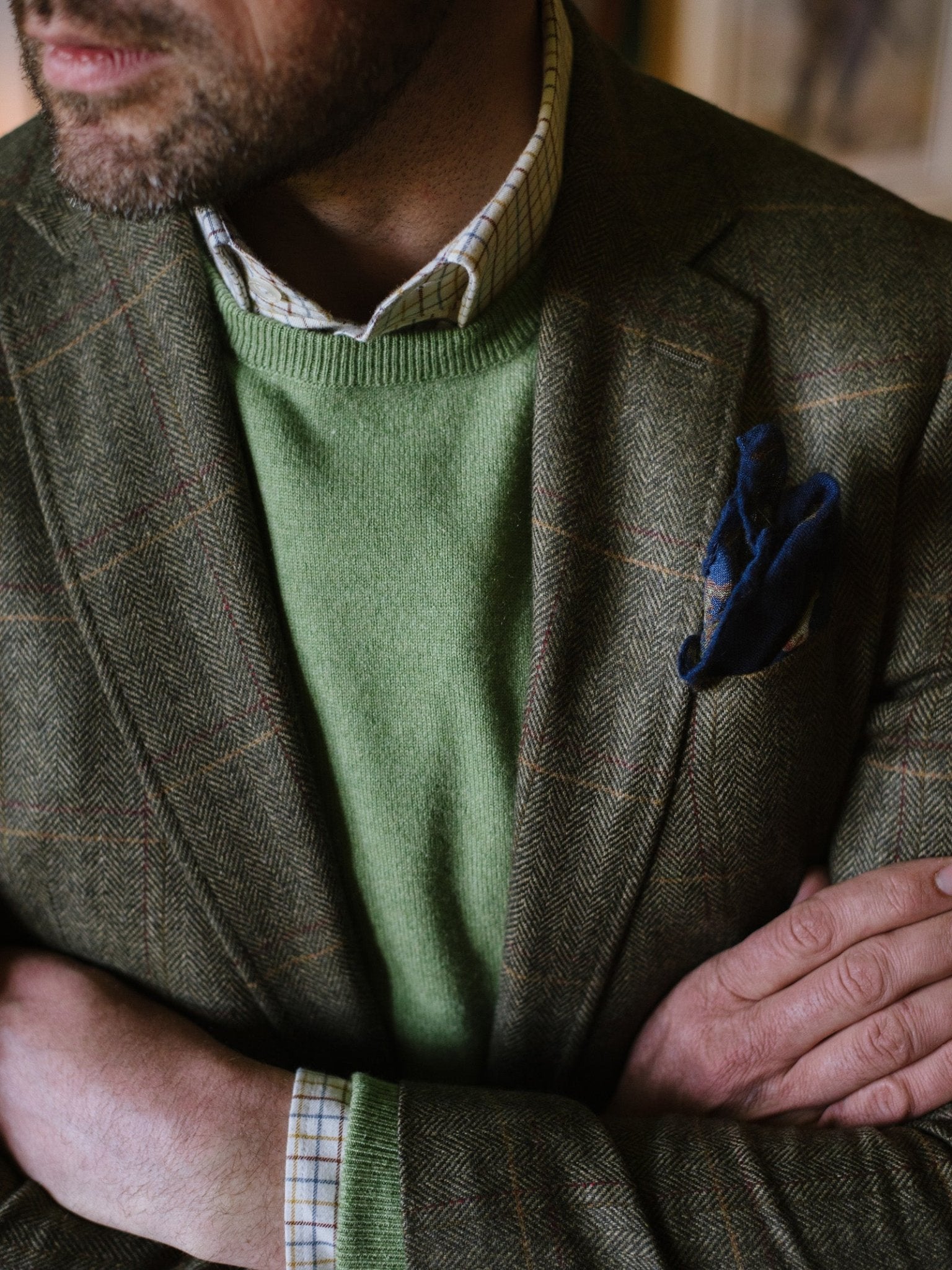 A man with crossed arms wears a Campbells of Beauly Cashmere Crew Neck Jumper in green over a checked shirt, layered with a brown plaid blazer and blue pocket square. His face is mostly out of frame.