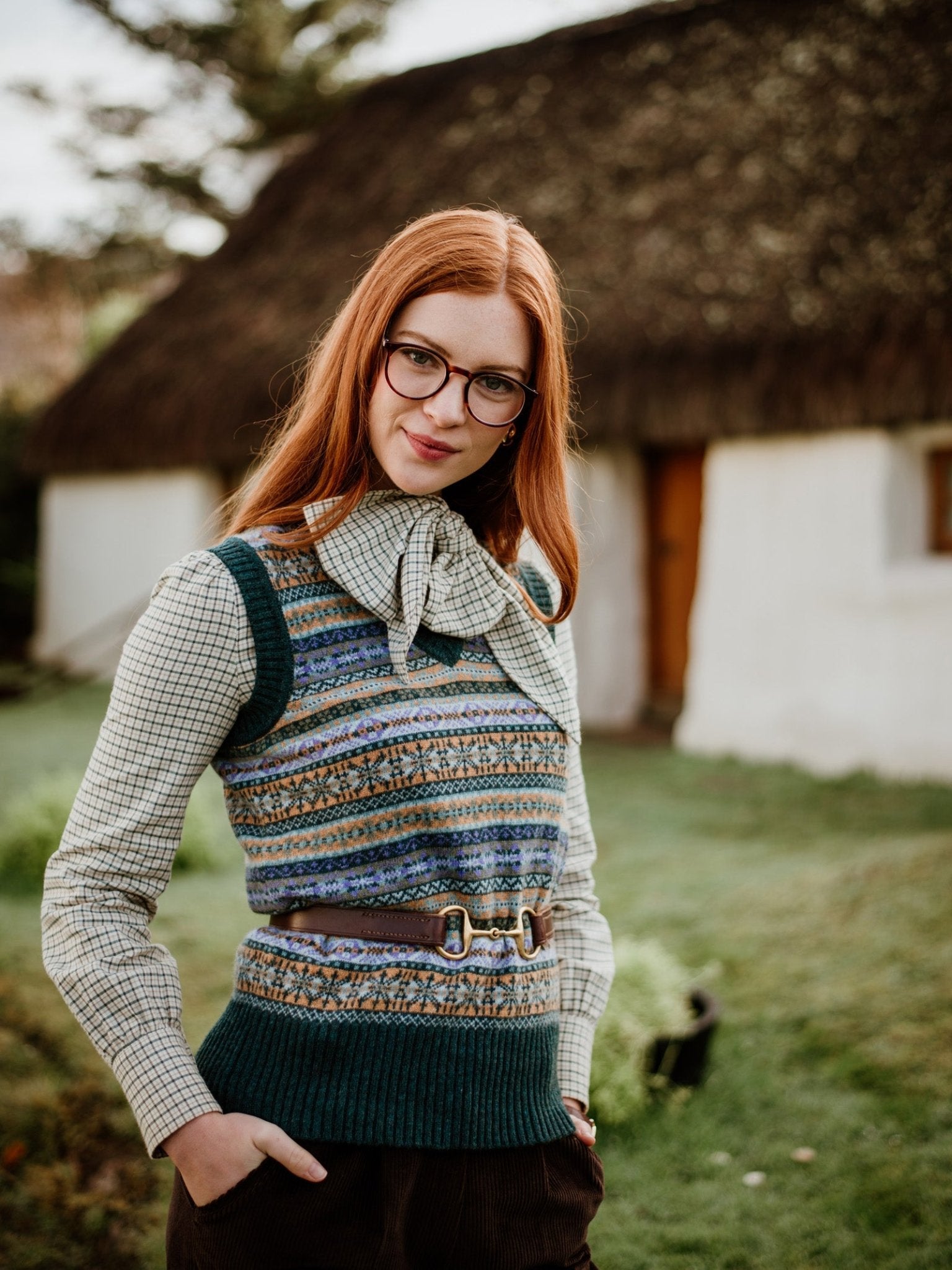 A woman with long red hair and glasses stands outside a white thatched cottage garden, wearing the Campbells of Beauly Cashmere V-Neck Fairisle Slipover over a checkered shirt with a necktie and belt.