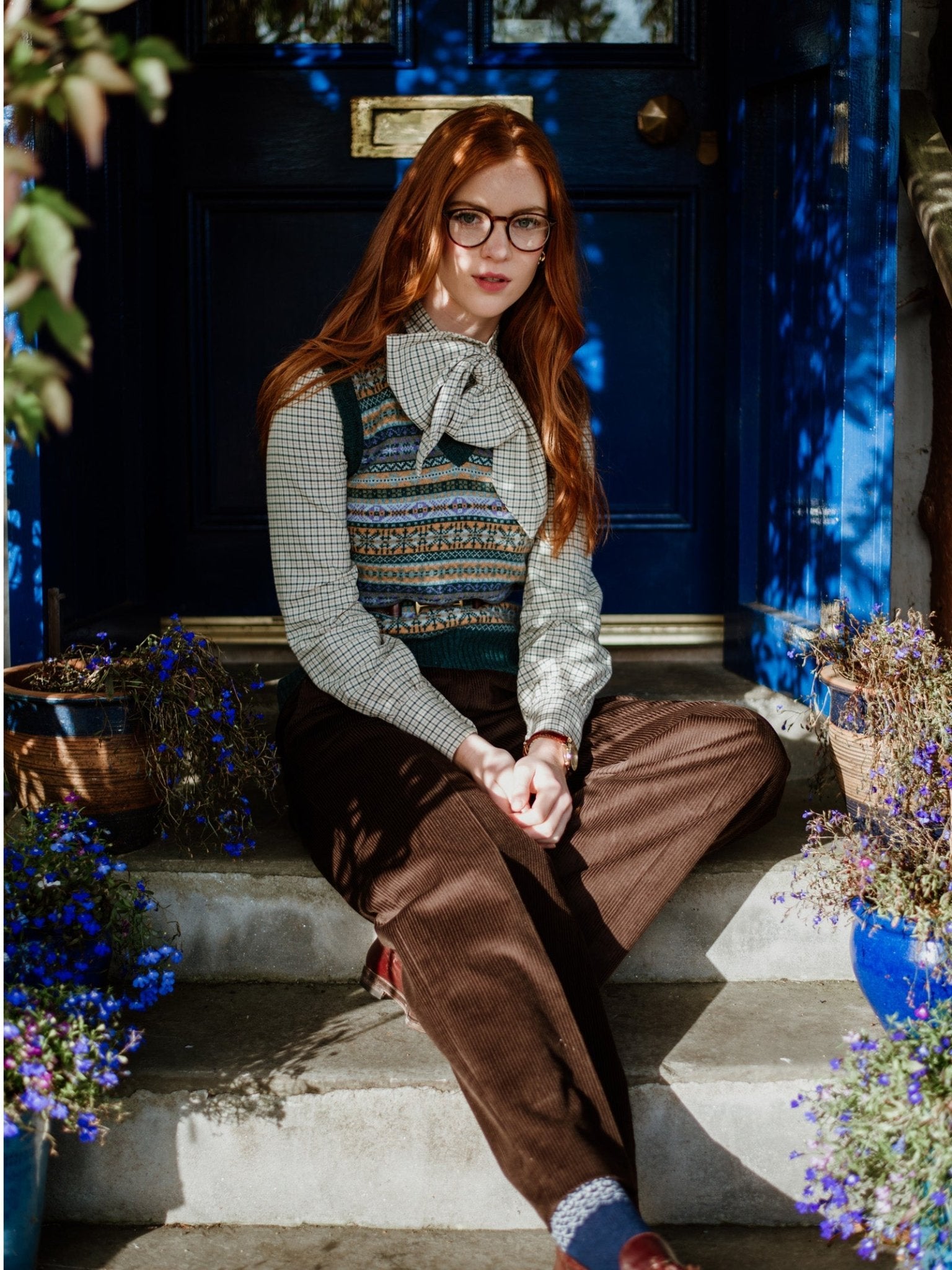 A young woman with long red hair and glasses sits on outdoor steps, wearing brown corduroy trousers and a Campbells of Beauly Cashmere V-Neck Fairisle Slipover with a traditional pattern over her checkered blouse. Blue flowers and a blue door are in the background.
