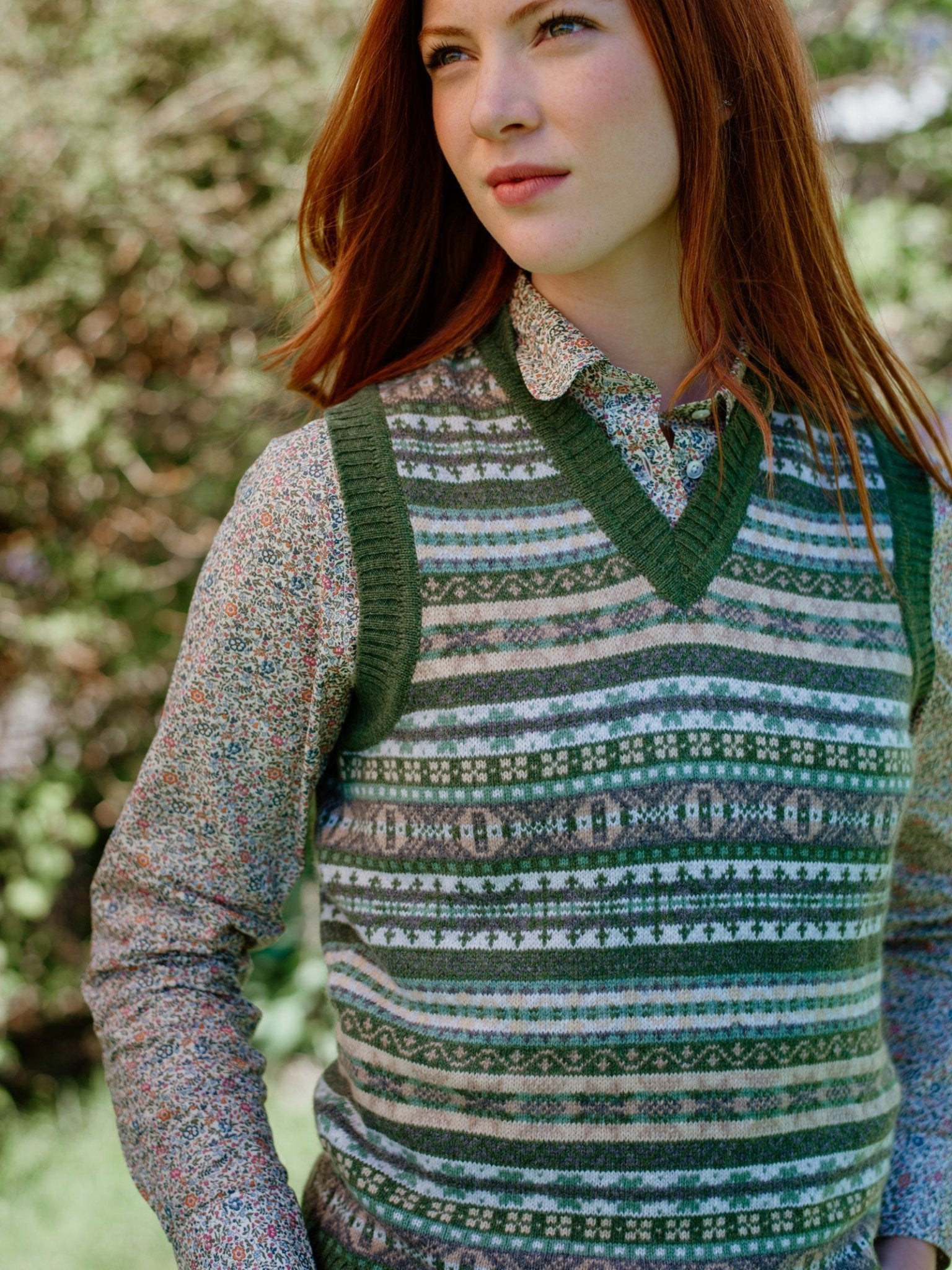 A young woman with long red hair stands outdoors wearing the Campbells of Beauly Cashmere V-Neck Fairisle Slipover over a floral long-sleeve shirt, looking away thoughtfully with blurred greenery behind her.