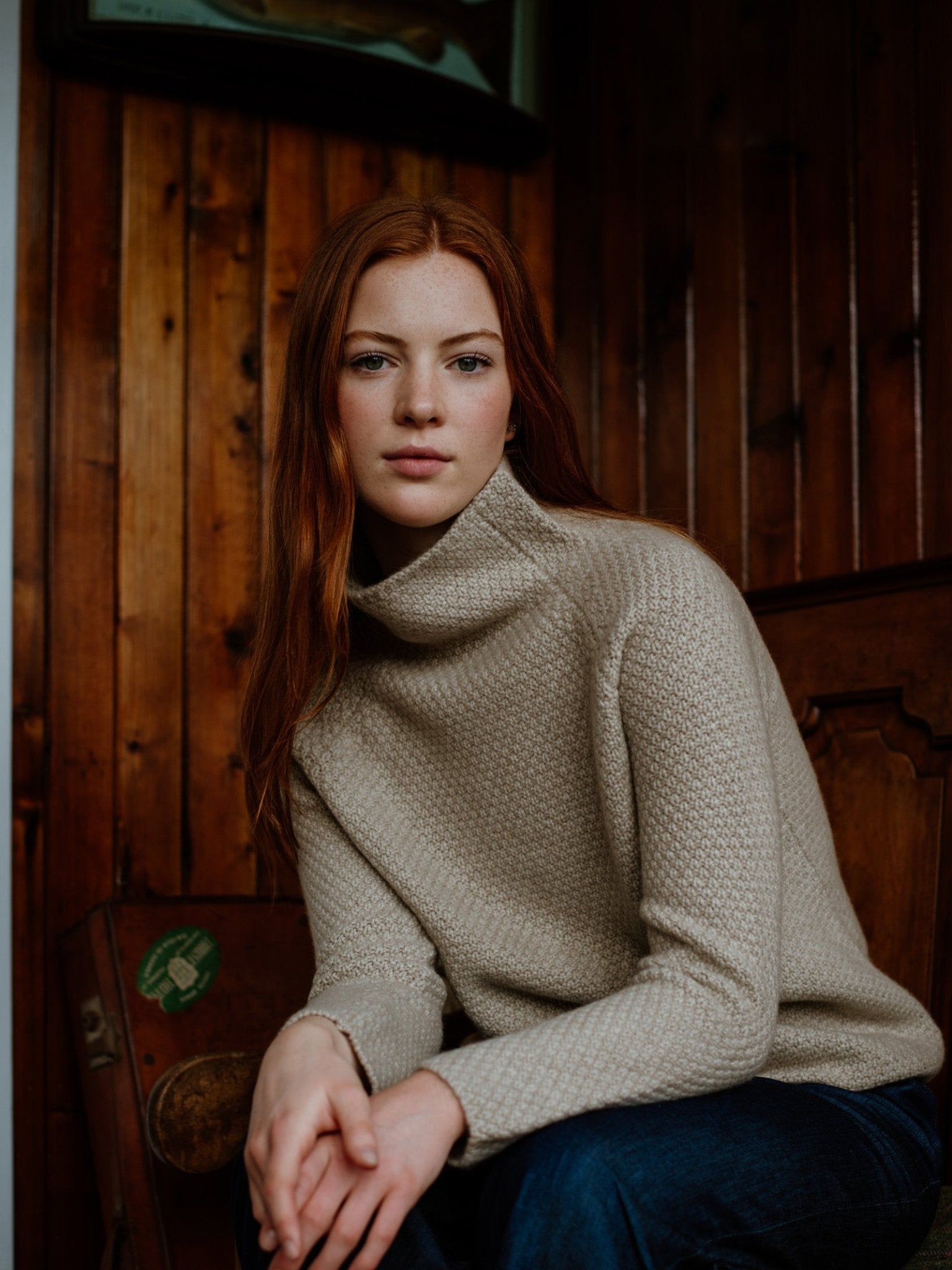 A young woman with long red hair sits indoors against a wooden wall, wearing Campbells of Beaulys Cashmere Funnel Neck Jumper in light beige and dark blue jeans, gazing calmly at the camera—a touch of Scottish Borders elegance.