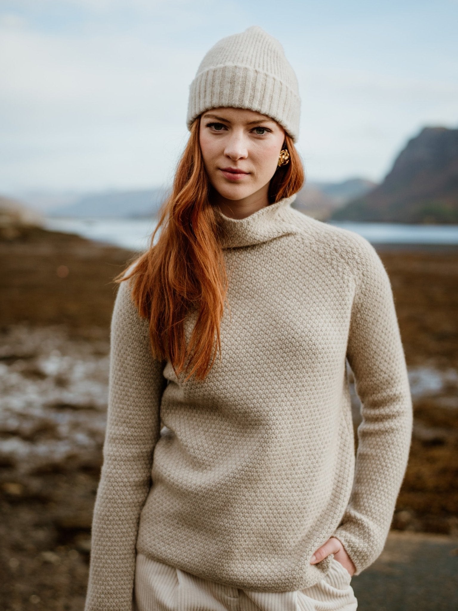 A woman with long red hair wears a beige Campbells of Beauly Cashmere Funnel Neck Jumper and matching beanie, standing outdoors by a rocky shoreline in the Scottish Borders, with mountains and water behind her.