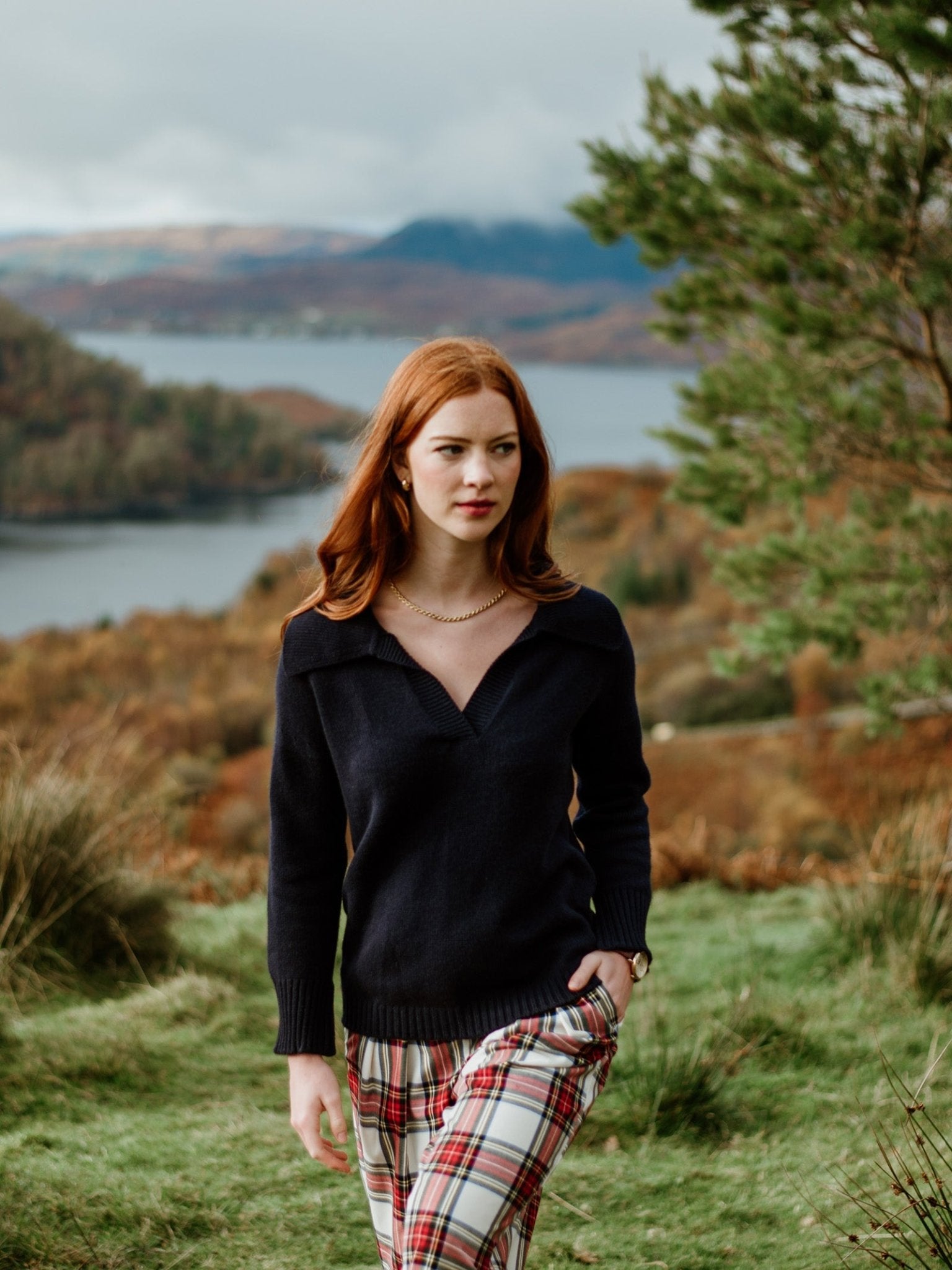 A young woman with long red hair wears a Campbells of Beauly Cashmere Shirt Jumper and plaid pants, standing outdoors in the Scottish Borders with a scenic lake and rolling hills in the background.