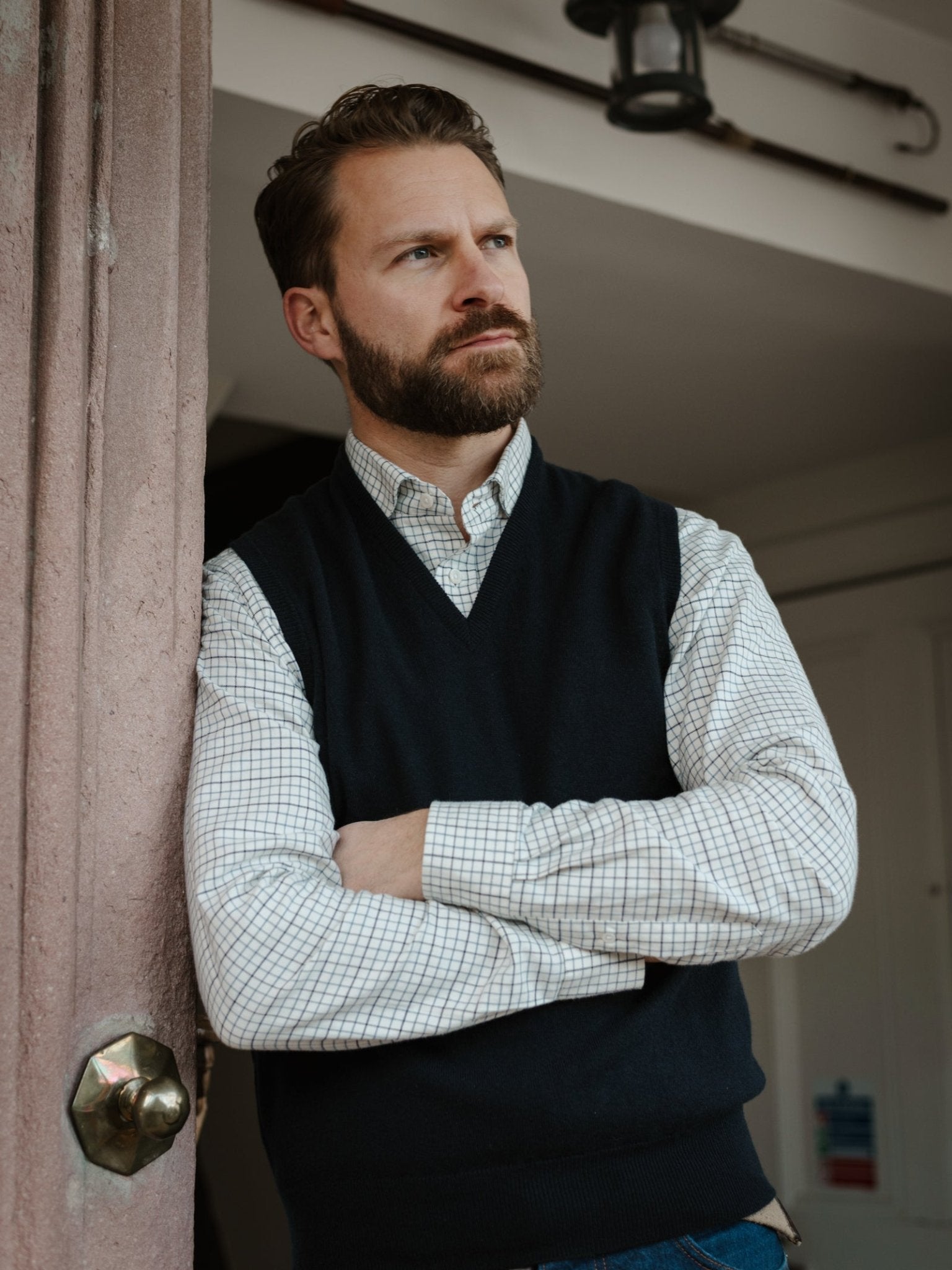A bearded man stands indoors, leaning against a doorframe with arms crossed, wearing a checkered shirt and the Cashmere Slipover by Campbell's of Beauly, made in Scotland, gazing thoughtfully into the distance.