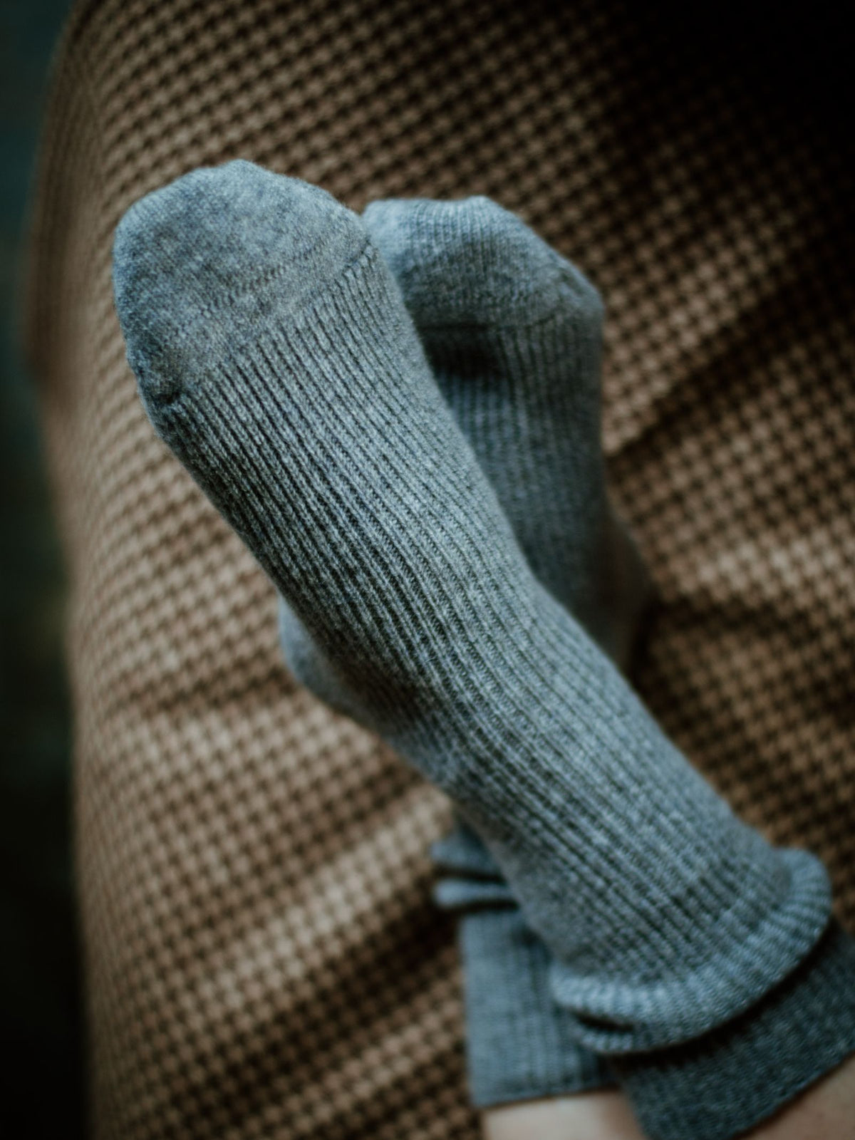 Close-up of feet in cozy Plain Cashmere Socks by Campbells of Beauly, legs crossed on a brown checkered surface—a luxurious gift inspired by the Scottish Borders.