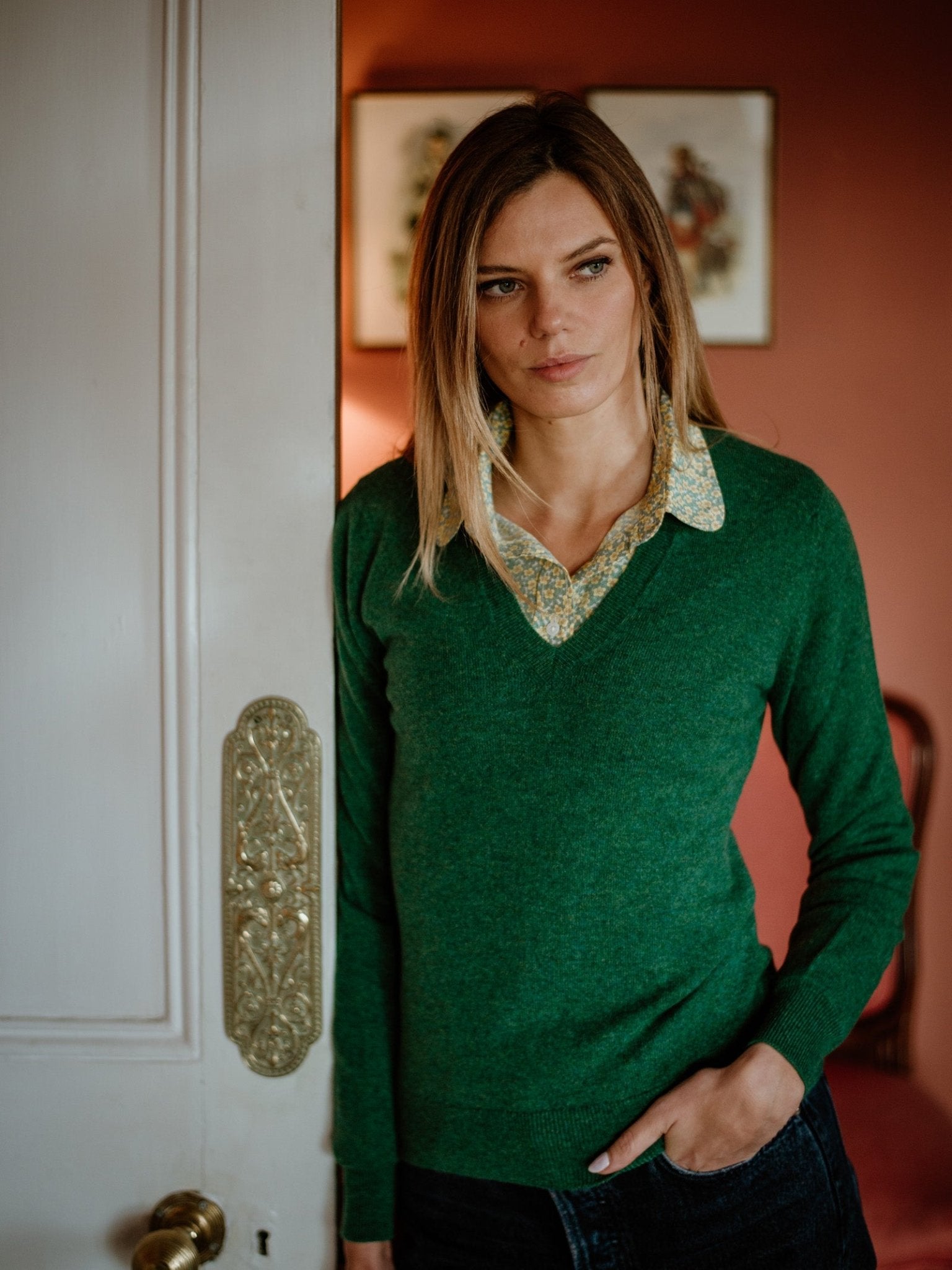 A woman with straight blond hair stands by an ornate white door, wearing a green Campbell’s of Beauly Cashmere V-Neck over a patterned collared shirt. Reddish walls and framed artwork are visible in the background.