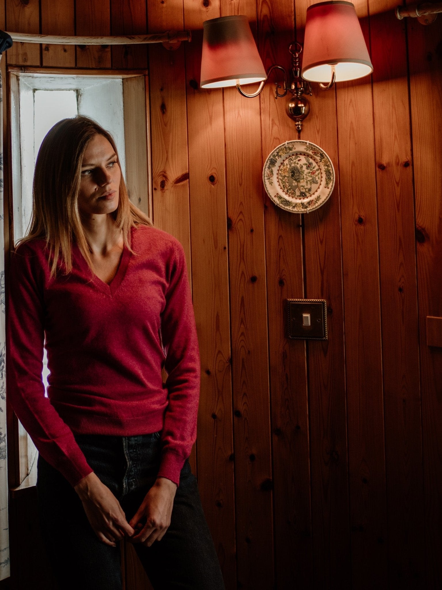 A woman wearing a Campbells of Beauly Cashmere V-Neck stands indoors by a wooden wall, gazing thoughtfully to the side. Light from a window and wall lamps brightens the space, with a decorative plate hanging behind her.