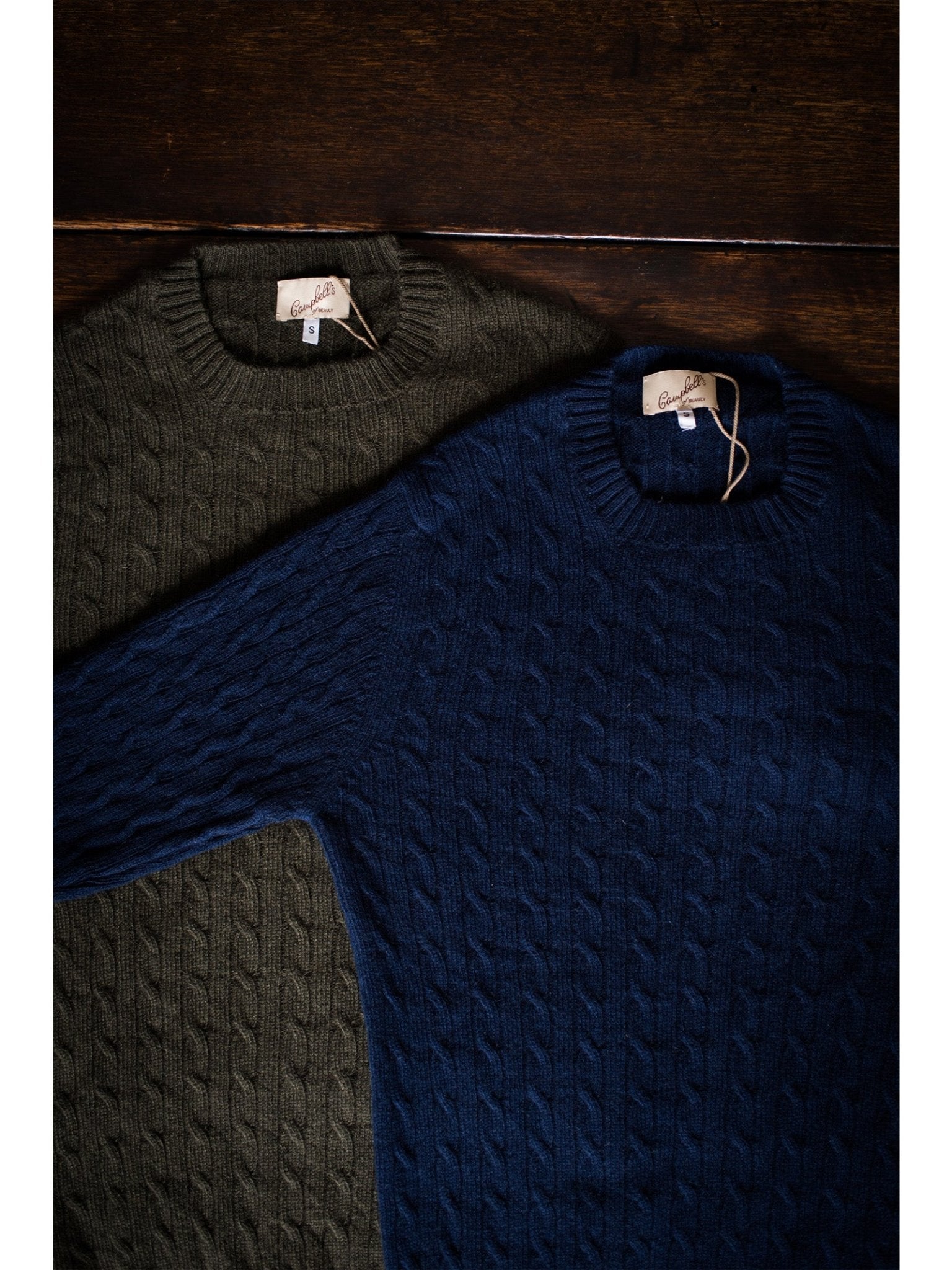Two Campbell's of Beauly Cashmere Cable Crew Jumpers—one olive green, one navy blue—lie on a dark wooden surface, each featuring a ribbed crew neck and a small tag at the neckline.