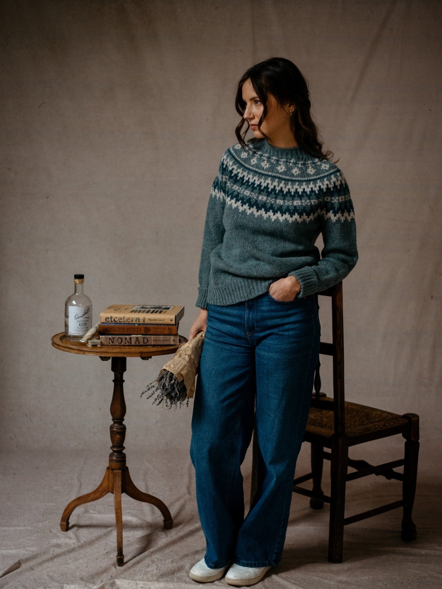 A woman wears the Chunky Fairisle Jumper by Campbells of Beauly with wide-leg jeans, standing by a wooden chair and small table with books, a scarf, and a bottle. She looks to the side and smiles against a neutral backdrop.