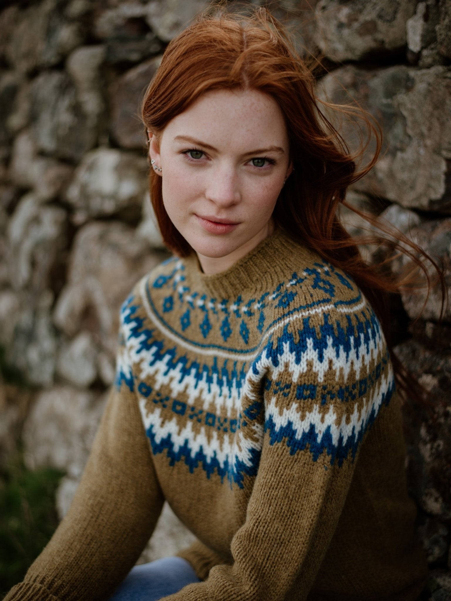 A woman with long red hair wears the Chunky Fairisle Jumper by Campbells of Beauly, a true winter staple, as she sits by a stone wall, gazing calmly at the camera.