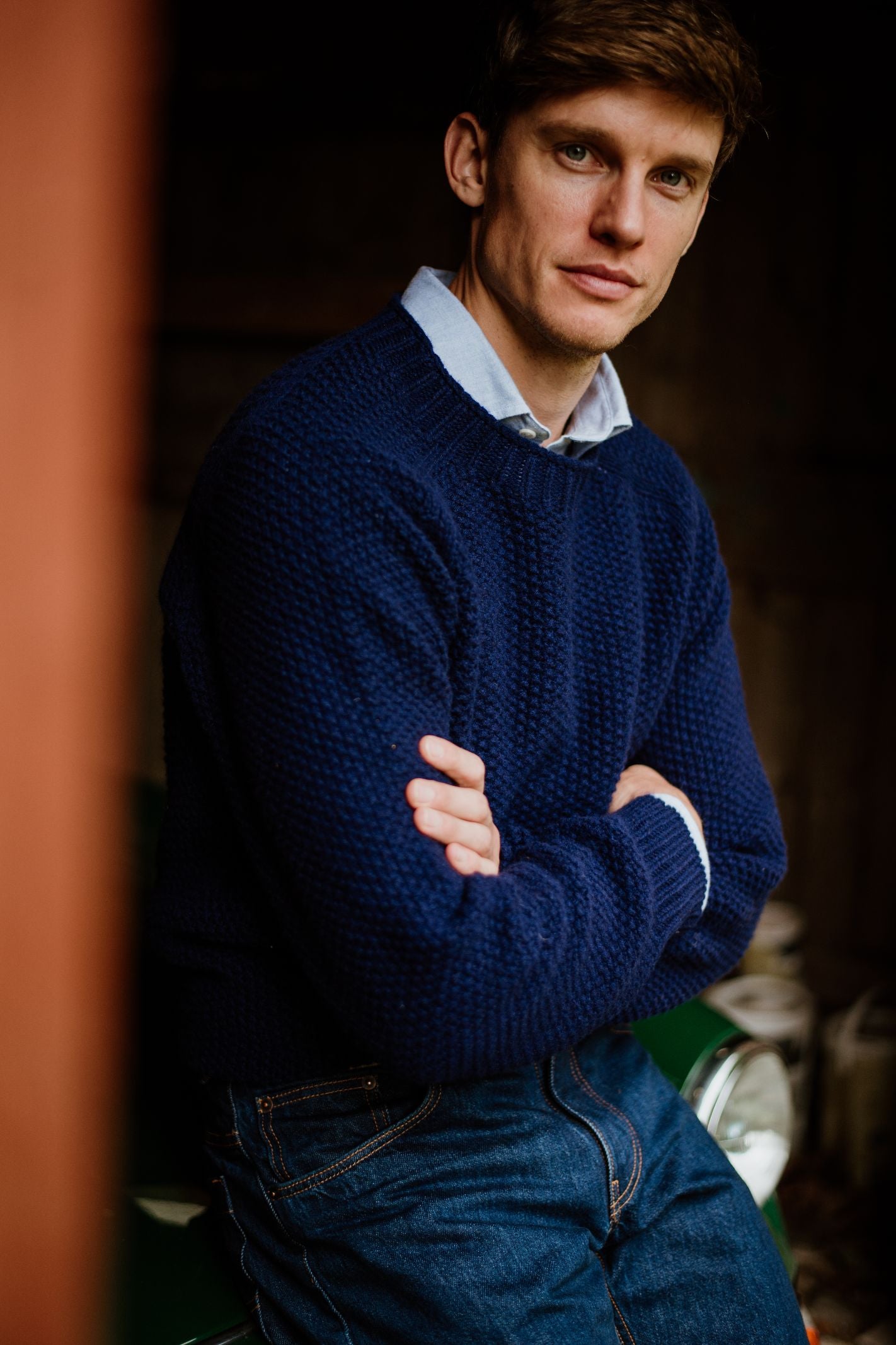 A man with light brown hair, wearing the Campbells of Beauly Geelong Lambswool Chunky Crew Jumper and jeans, sits with his arms crossed, looking at the camera against a dim, softly blurred background.