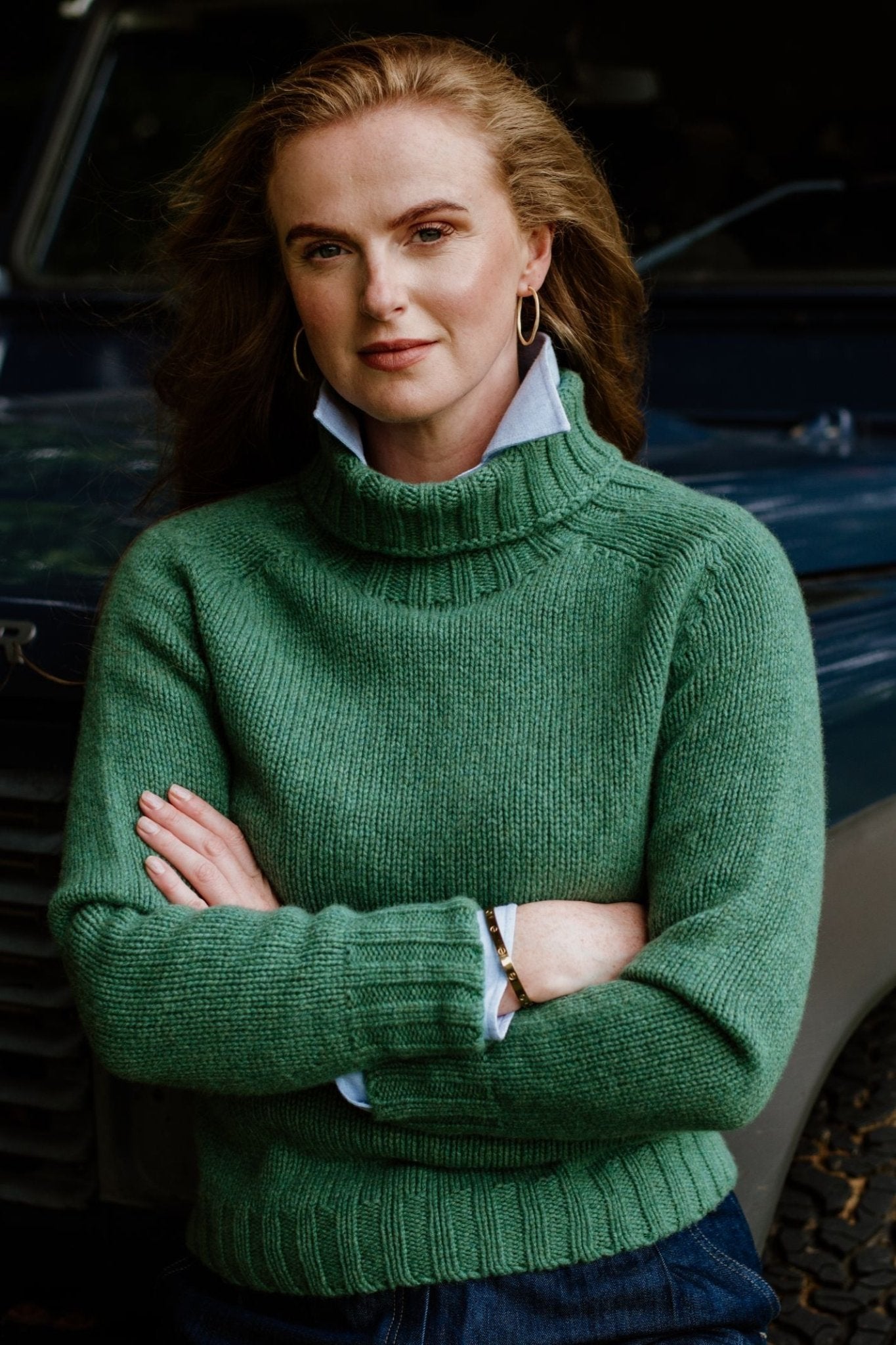 A woman stands confidently in front of a car, arms crossed, wearing the Campbells of Beauly Chunky Roll Neck Sweater in green over a collared shirt. Her long, wavy hair completes the stylish look.
