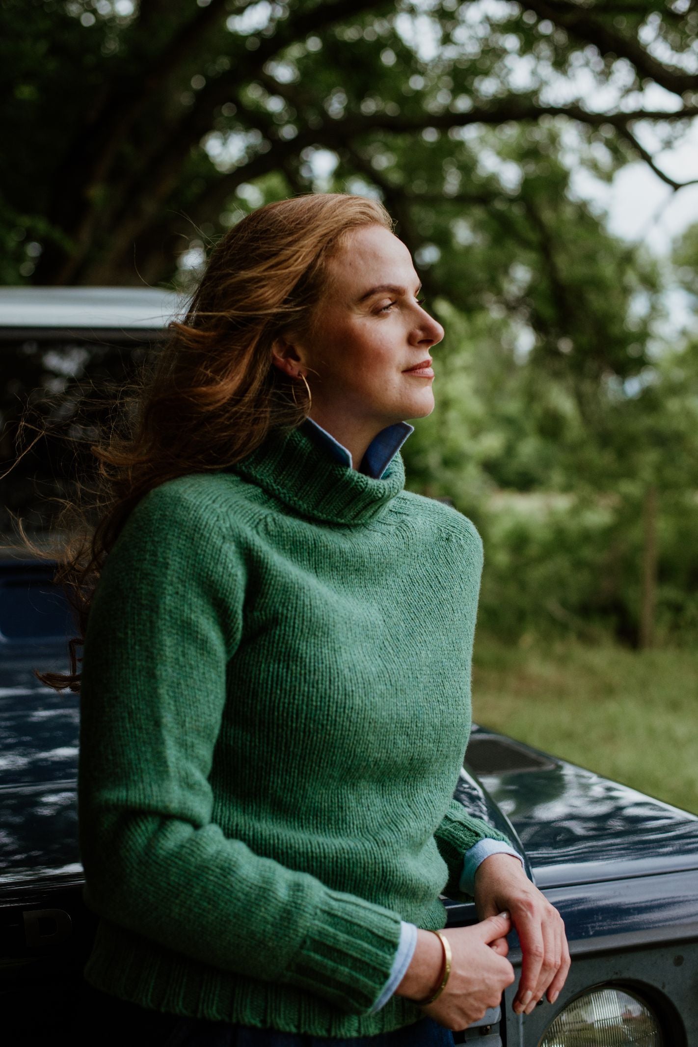 A woman with long brown hair stands outdoors beside a vehicle, eyes closed in calm, wearing the Campbell’s of Beauly Chunky Roll Neck Sweater. Lush green trees fill the background.