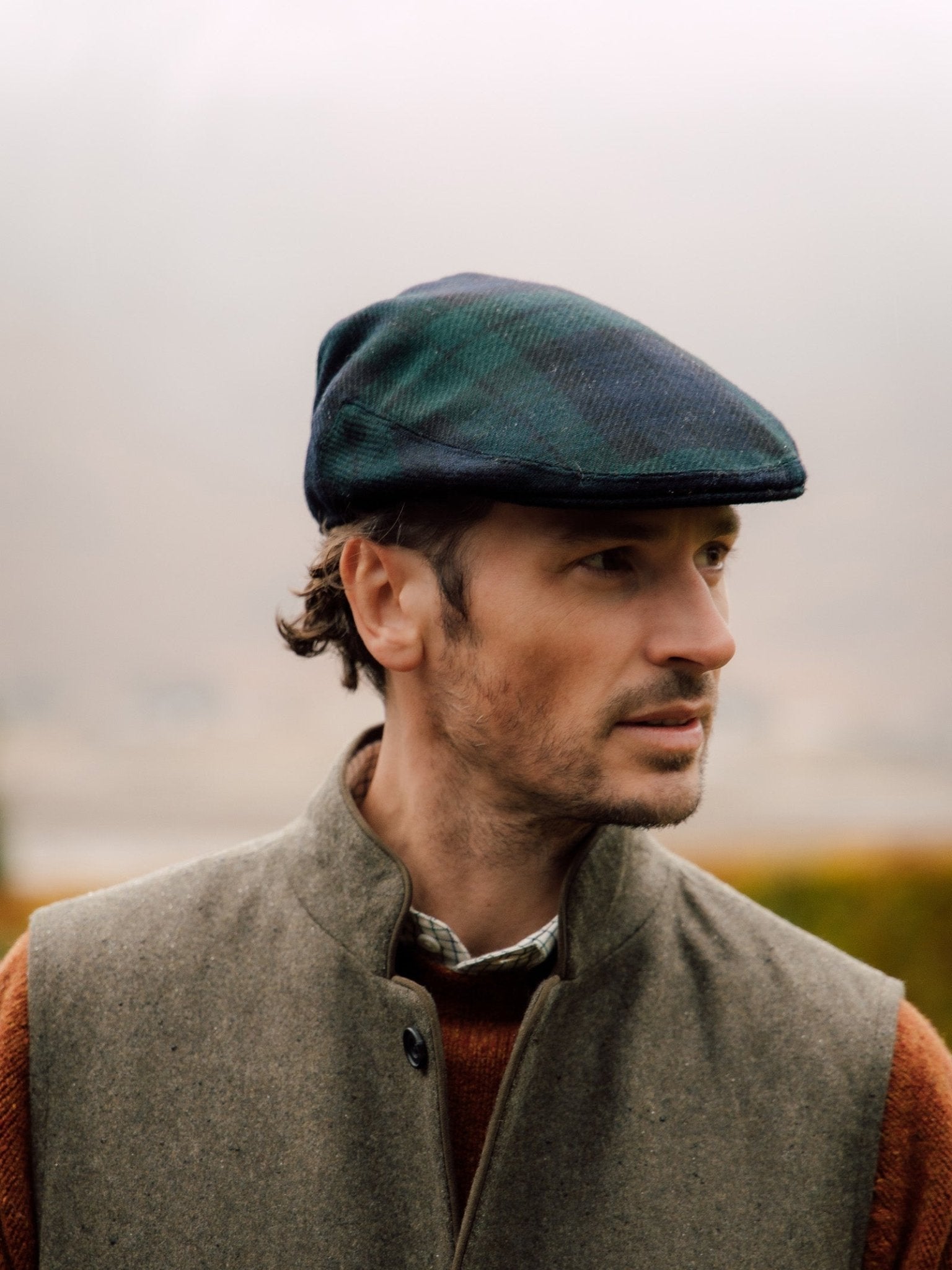 A man wears a Campbells of Beauly Blackwatch Tartan Conon Cap and a gray tweed vest over a brown sweater, gazing to the side outdoors against a softly blurred background.