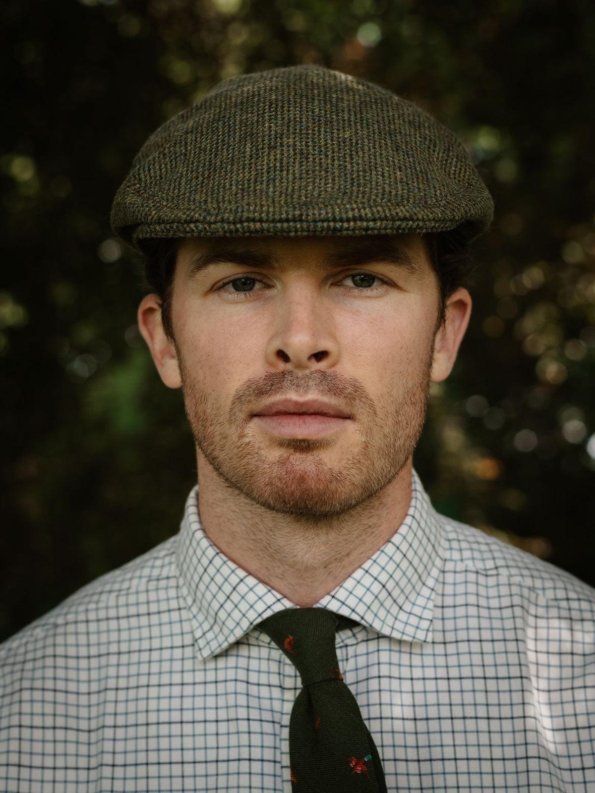 A man outdoors in a checkered shirt and green patterned tie wears the Conon Cap by Campbell’s of Beauly. The greenery in the background is blurred, highlighting his neutral expression.