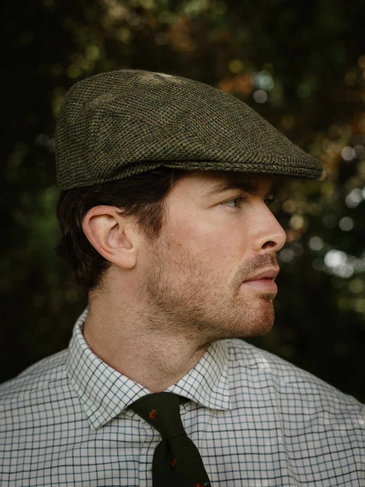 A man in a green Conon Cap by Campbell’s of Beauly, with a checked shirt and green tie, looks to the side against a blurred green background, evoking classic British style.