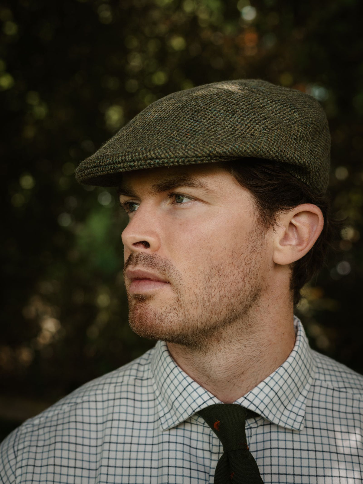 A man wears the Conon Cap by Campbell’s of Beauly, paired with a checkered shirt and green tie. He looks to the side with a neutral expression, set against blurred foliage.