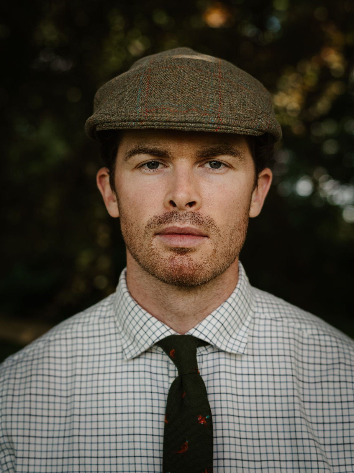A man wearing the Conon Cap by Campbell’s of Beauly, a brown British tweed cap, stands outdoors in a checked shirt and green tie, looking directly at the camera with a neutral expression.