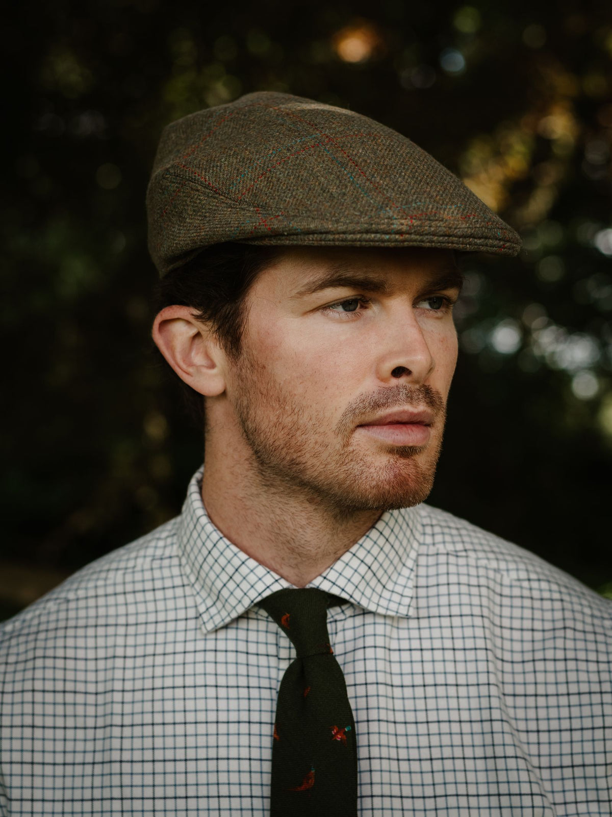A man wearing the Conon Cap by Campbell’s of Beauly, a checkered shirt, and a green tie stands outdoors, gazing to the side with a neutral expression.