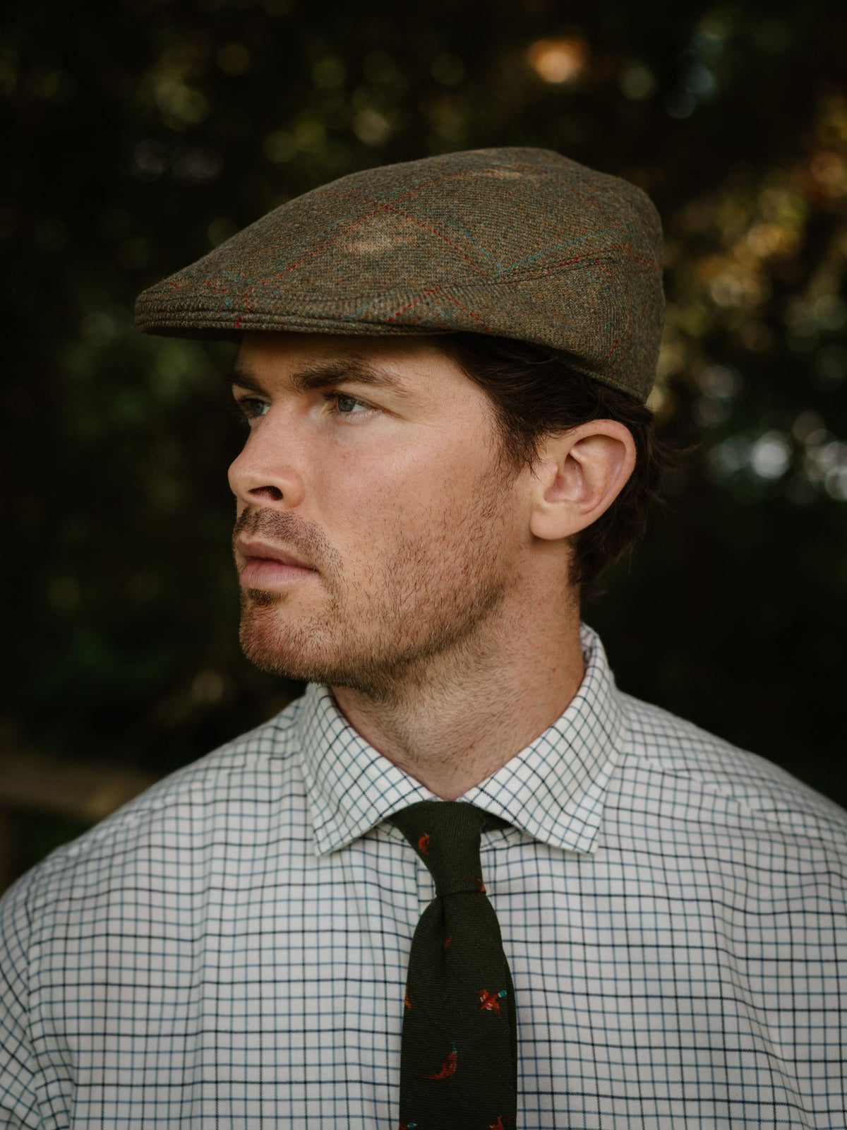 A man in a checkered shirt, patterned tie, and Campbell’s of Beauly Conon Cap gazes sideways outdoors against a softly blurred green background.