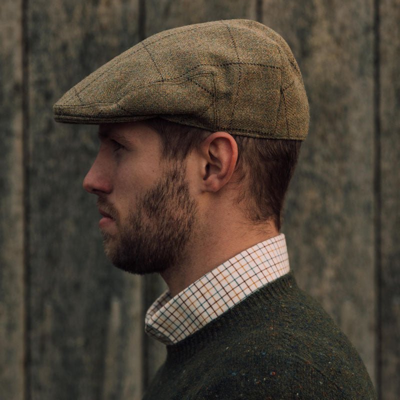 A bearded man in profile, wearing the Campbell's of Beauly Auld Stock Conon Cap, a green sweater, and a checkered collared shirt, stands against a wooden background.