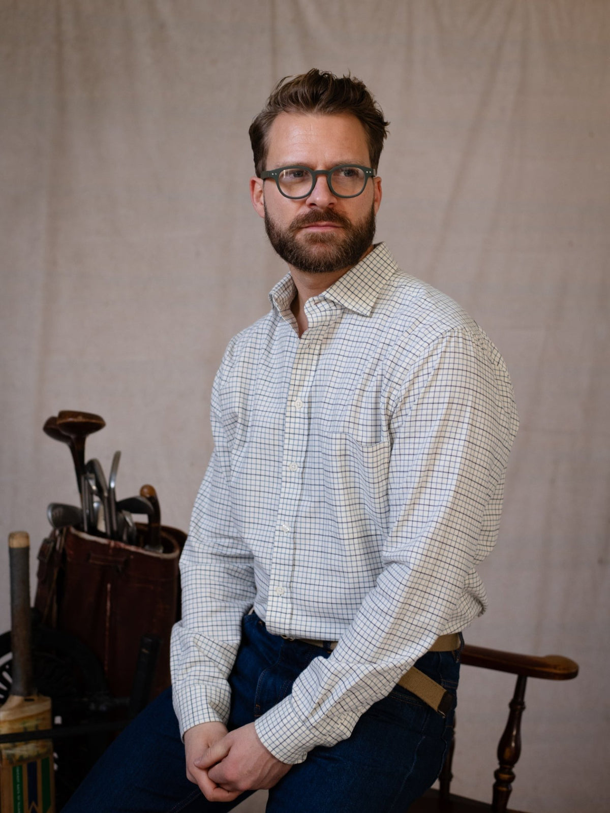 A man with glasses and a beard, wearing the Campbell’s of Beauly Windowpane Shirt in Windowpane Blue Check and a pair of jeans, sits on a chair. In the background, against a plain light-colored backdrop, there is a leather bag with tools and some wooden objects.