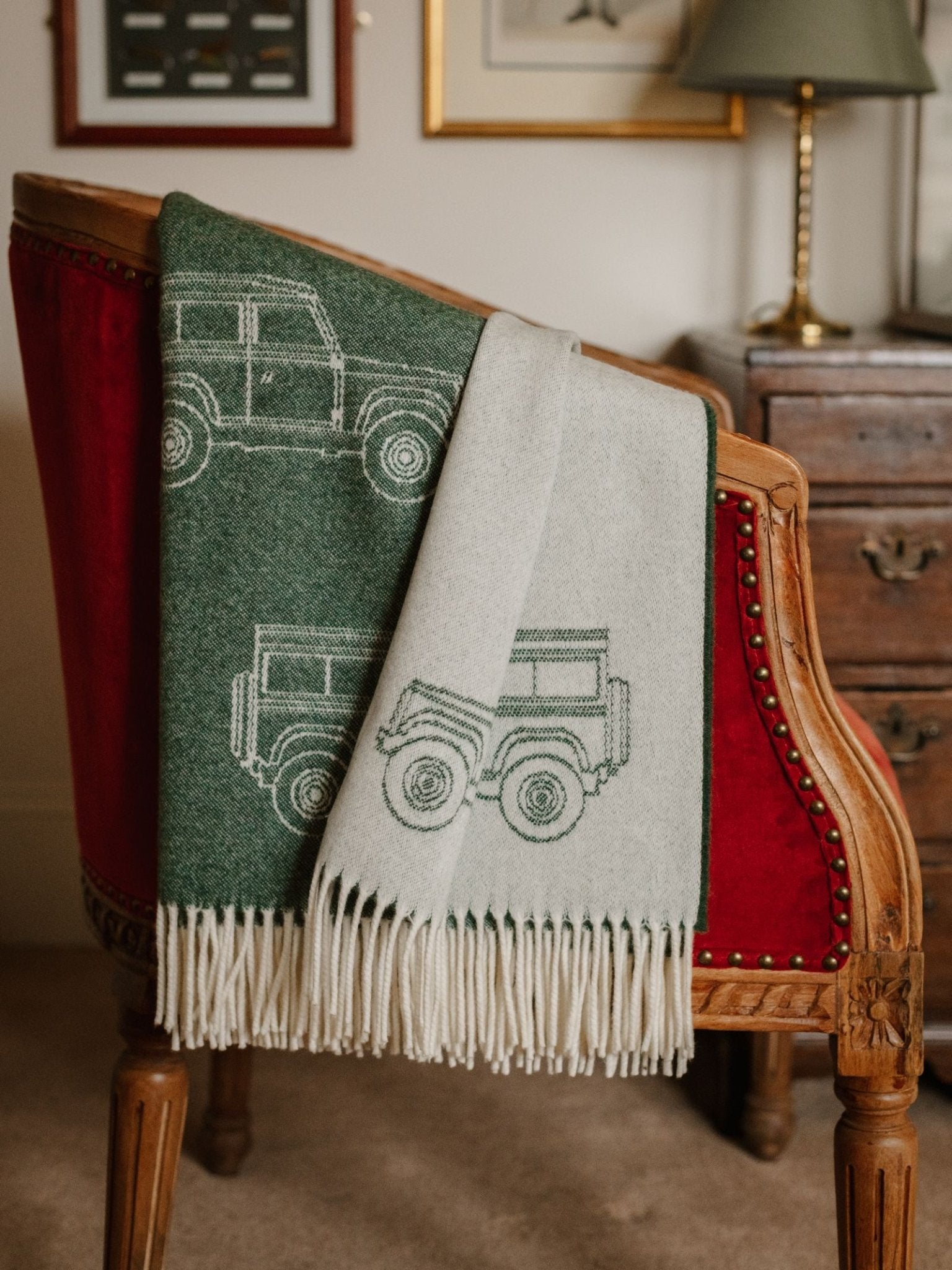 The Campbell's of Beauly Land Rover Defender Jacquard Throw, featuring green and white SUV line drawings, is draped over a vintage red velvet chair beside a wooden dresser in a cozy room.
