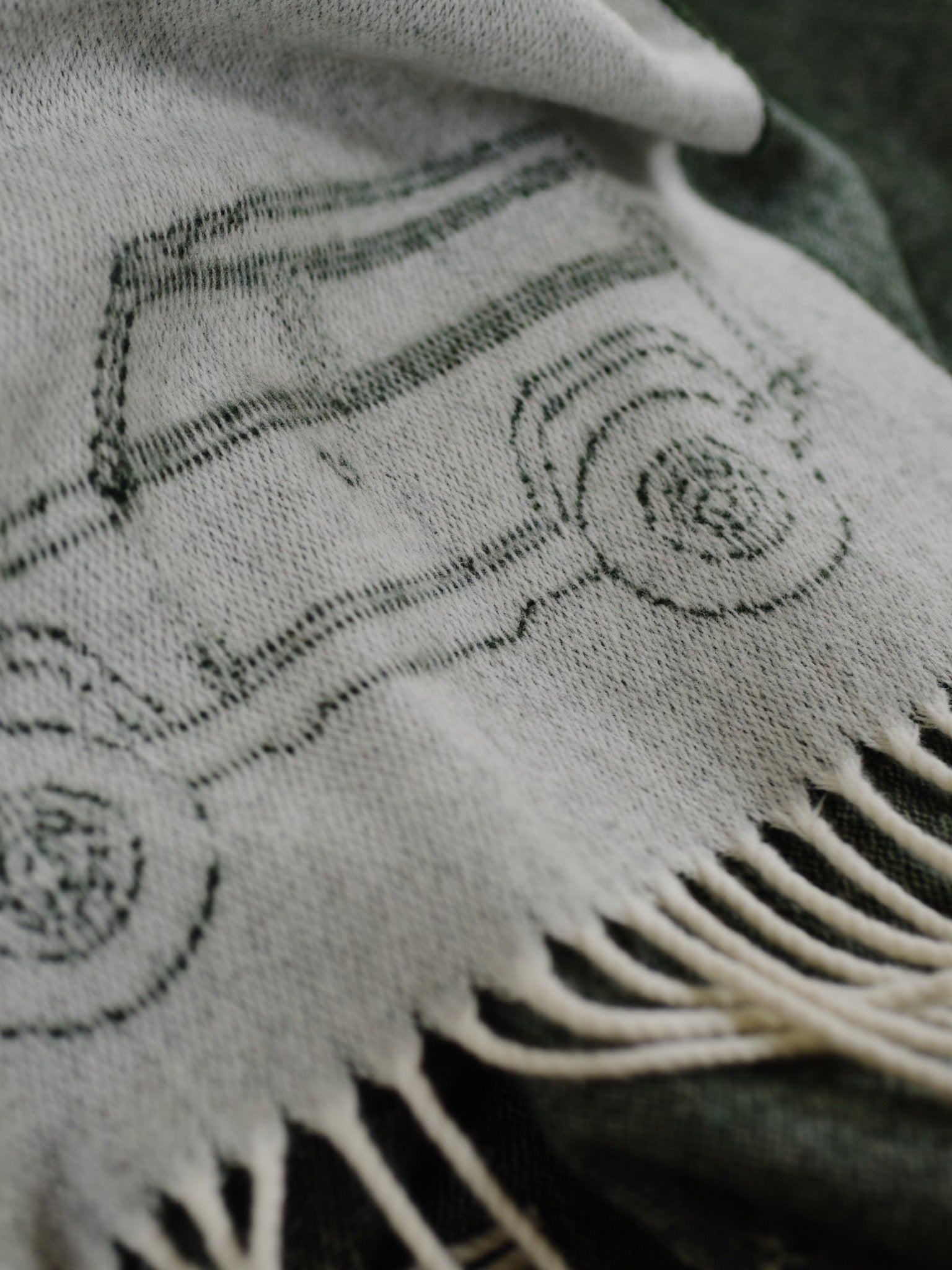 The Land Rover Defender Jacquard Throw by Campbells of Beauly is a cream wool blanket with a green Defender line drawing and fringed edges, shown atop a dark green textile background.