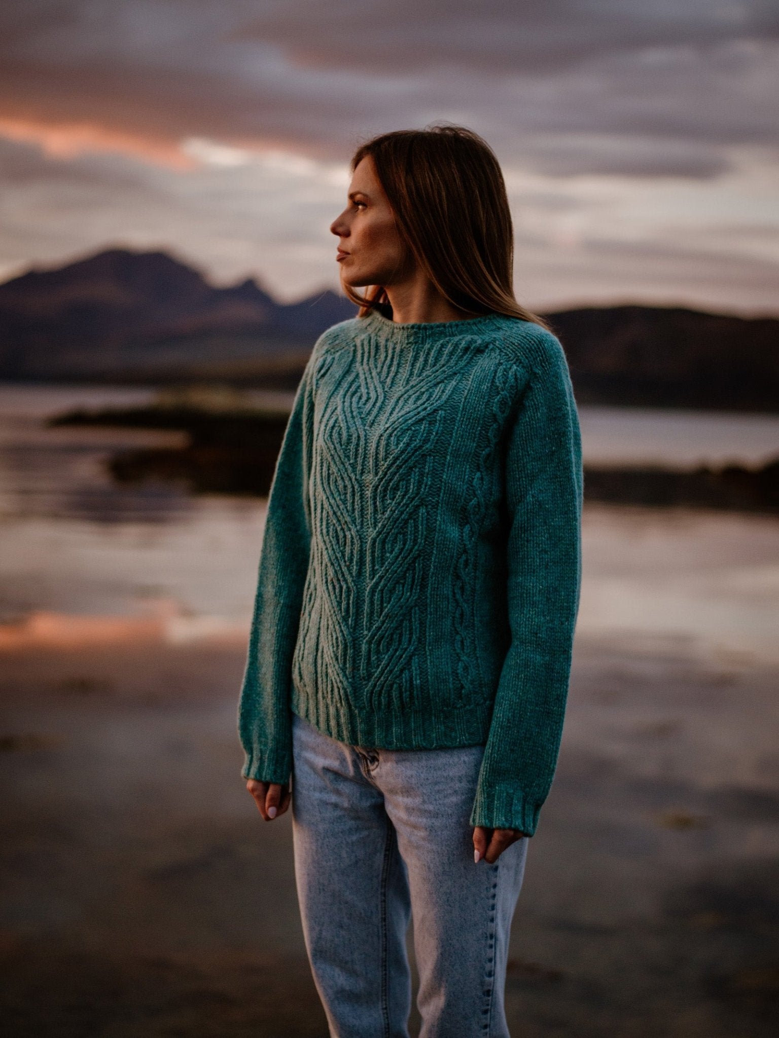 A woman in a teal Donegal Lambswool Cable Crew Neck Jumper by Campbells of Beauly and light jeans stands beside a calm lake at sunset, mountains and clouds behind her, looking to her left.