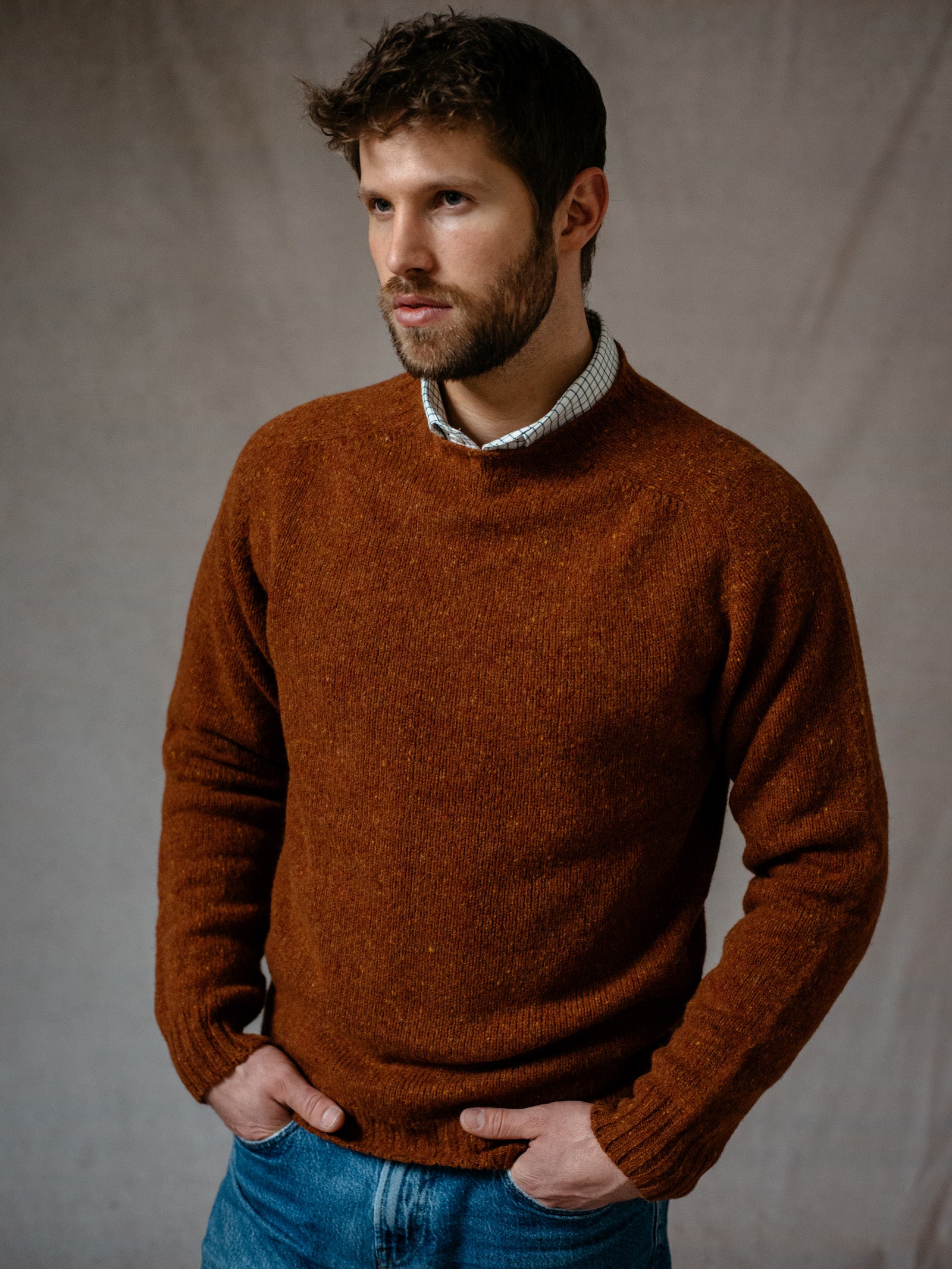 A bearded man with short brown hair stands against a plain background, wearing a rust Donegal Lambswool Crew Neck Jumper by Campbells of Beauly. The flecked texture adds character as he gazes to the side, hands in his jeans pockets.