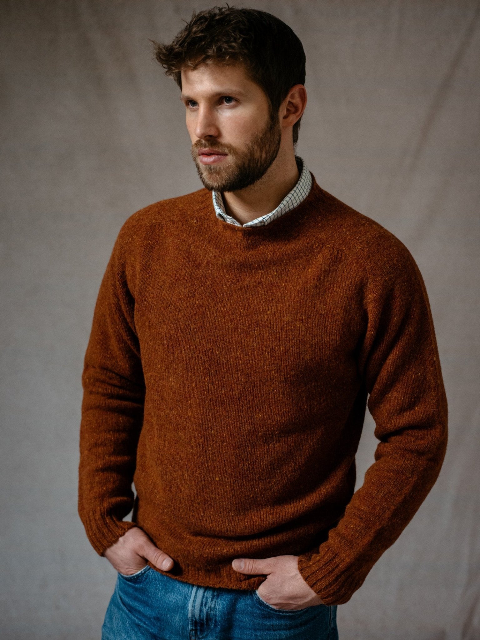 A man with short brown hair and a beard wears a rust Donegal Lambswool Crew Neck Jumper by Campbells of Beauly over a collared shirt and blue jeans, standing with hands in his pockets against a neutral textured background.