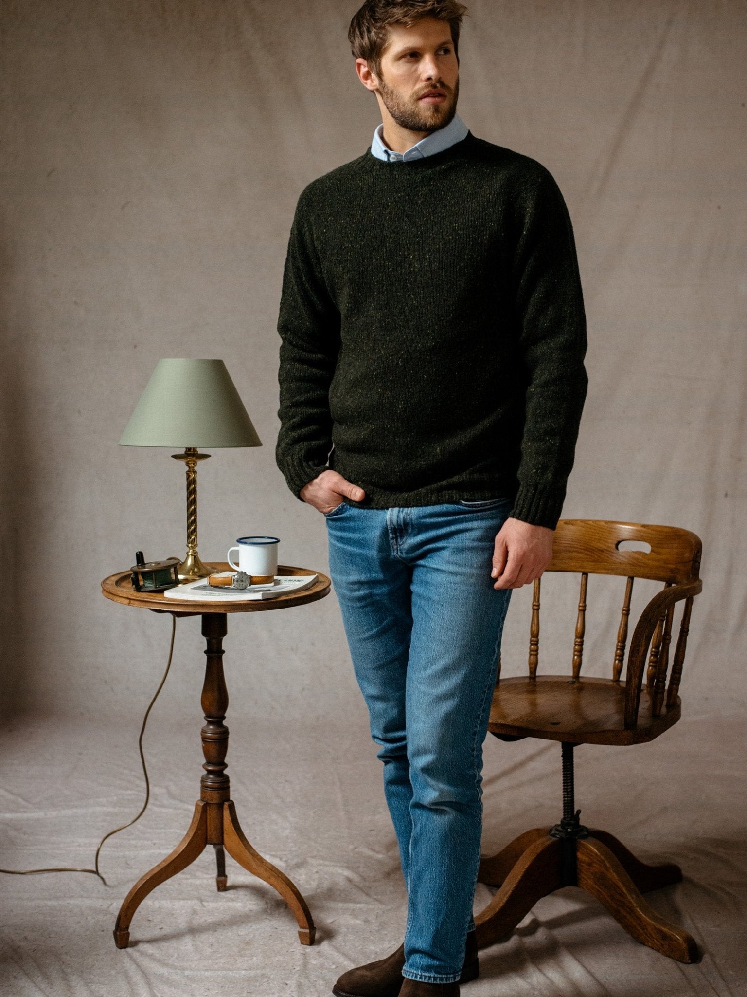 A man with short brown hair and a beard stands indoors by a small round wooden table, wearing a dark green Campbells of Beauly Donegal Lambswool Crew Neck Jumper over a collared shirt, blue jeans, and brown shoes.