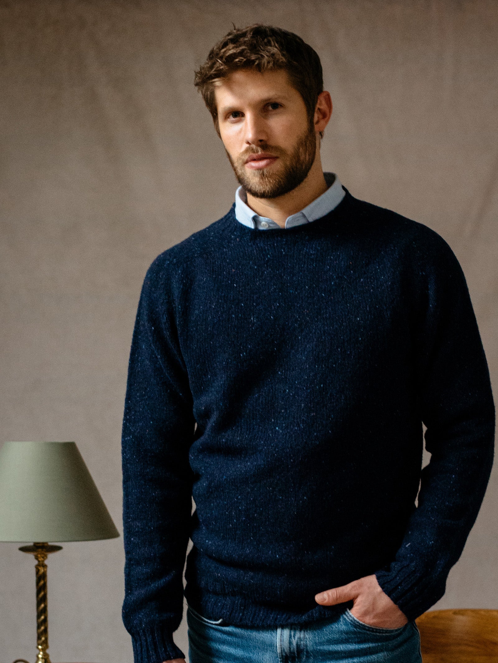 A man with short brown hair and a beard wears a dark blue Donegal Lambswool Crew Neck Jumper by Campbells of Beauly, its flecked texture over a light blue collared shirt and jeans. He stands indoors near a gold lamp with a green shade against a neutral background.