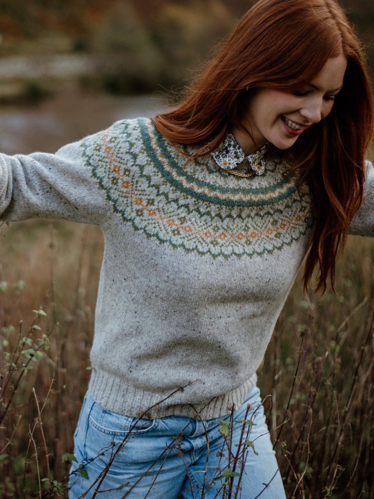 A young woman with long red hair smiles outdoors in tall grass, wearing the Campbells of Beauly Donegal Lambswool Fairisle Crew sweater over a floral-collared shirt and light blue jeans.