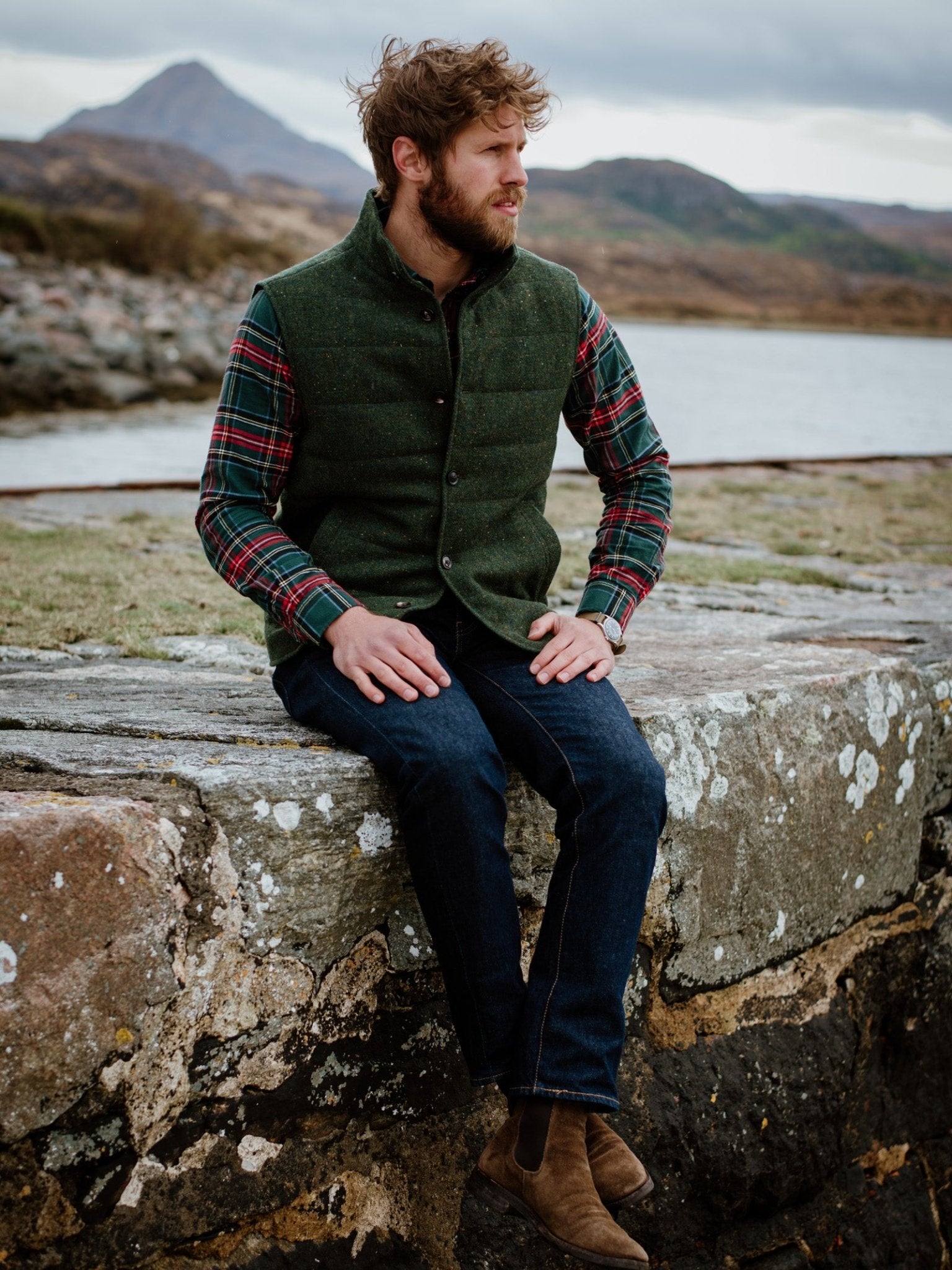 A bearded man sits on a stone wall by a lake, hills and clouds behind him, wearing Campbells of Beauly Donegal Lambswool Green Quilted Gilet over a plaid shirt, blue jeans, and brown boots—a classic modern country style.