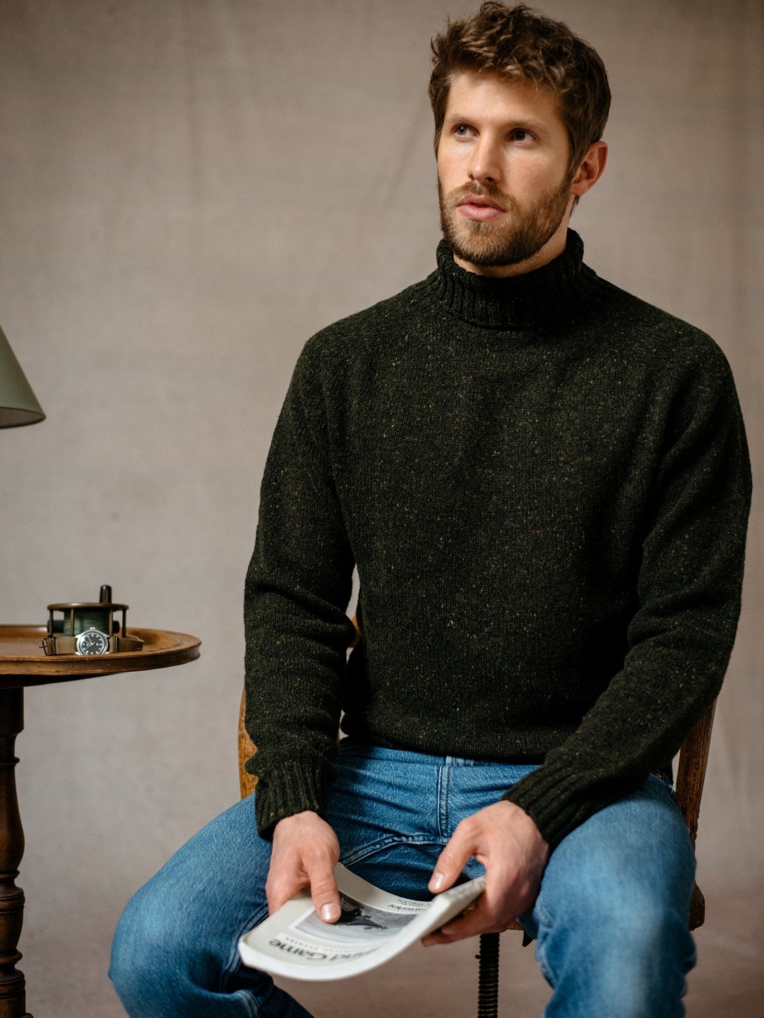 A man with short brown hair and a beard, wearing a Campbells of Beauly Donegal Lambswool Poloneck in dark green and blue jeans, sits on a chair holding a newspaper. A table with a watch and lamp is beside him as he gazes thoughtfully ahead.