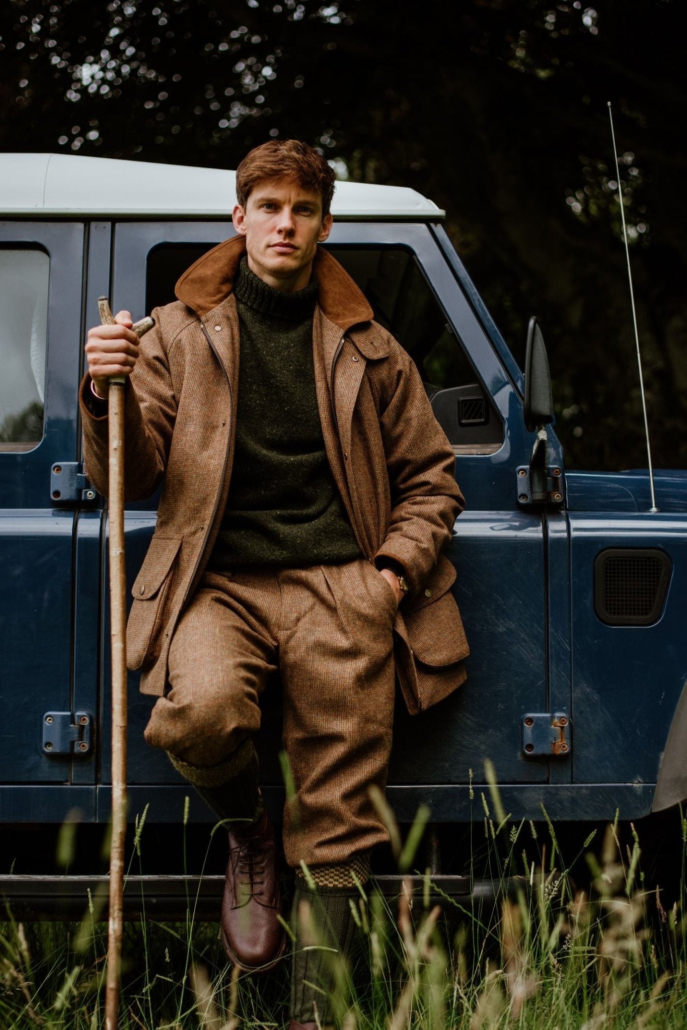 A man wearing the Campbells of Beauly Donegal Lambswool Poloneck in brown leans against a blue vintage vehicle, holding a wooden walking stick. He stands on grass with trees behind him, looking directly at the camera.