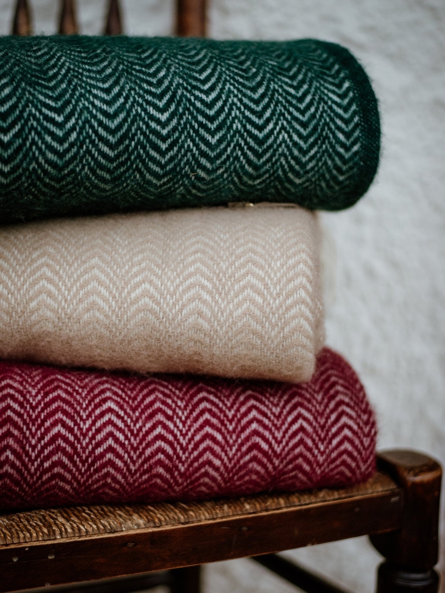 Three folded Dune Throws by Campbells of Beauly, with a chevron pattern in dark green, cream, and burgundy, are stacked on a wooden chair with a woven seat—a cozy addition inspired by Scottish weaving against a white textured wall.