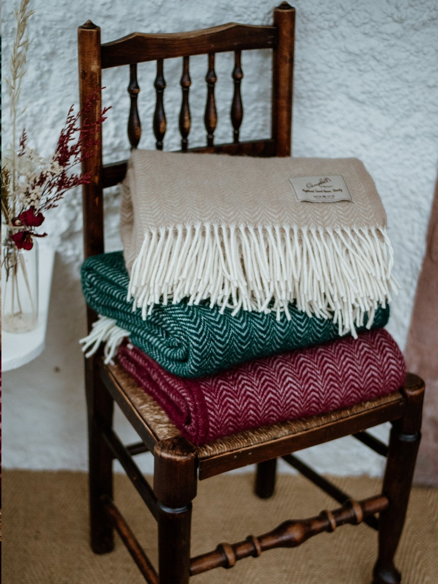 A wooden chair holds three folded, fringed Campbells of Beauly Dune Throws in beige, green, and burgundy lambswool. Nearby, dried flowers add a cozy, rustic touch—perfect for enhancing your home.