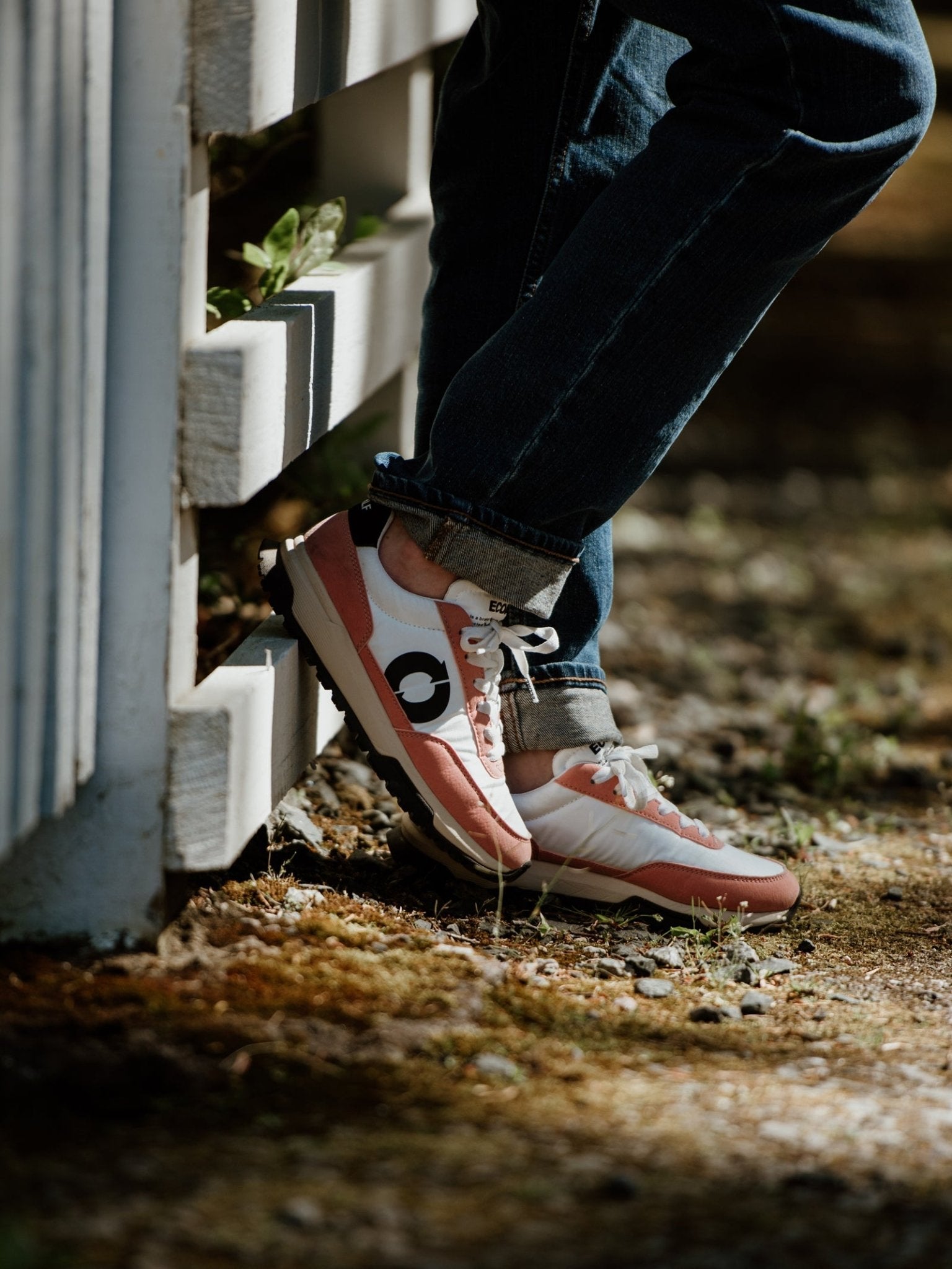 A person wearing jeans and ECOALF Ventura Trainers in pink, made with recycled polyester, stands outdoors leaning against a white wooden fence with one foot on the fence as sunlight casts soft shadows on the ground.