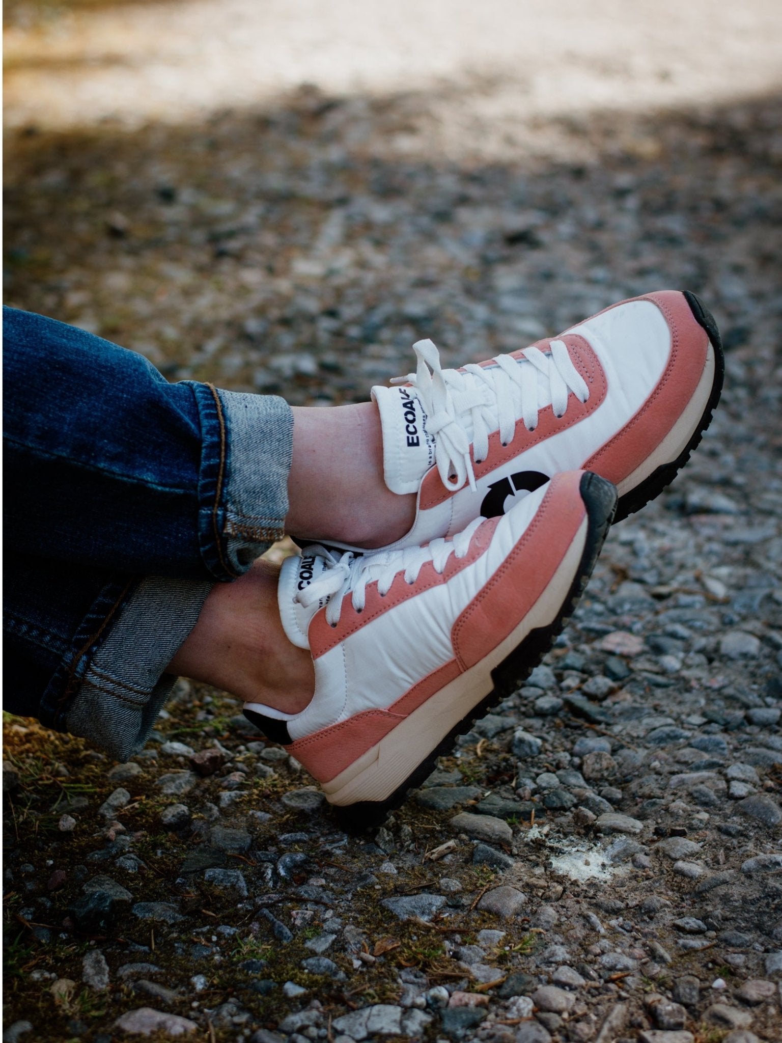 A person wearing cuffed blue jeans and ECOALF Ventura Trainers—white sneakers with pink accents made from recycled polyester—sits outdoors on a gravel path, legs crossed at the ankles.