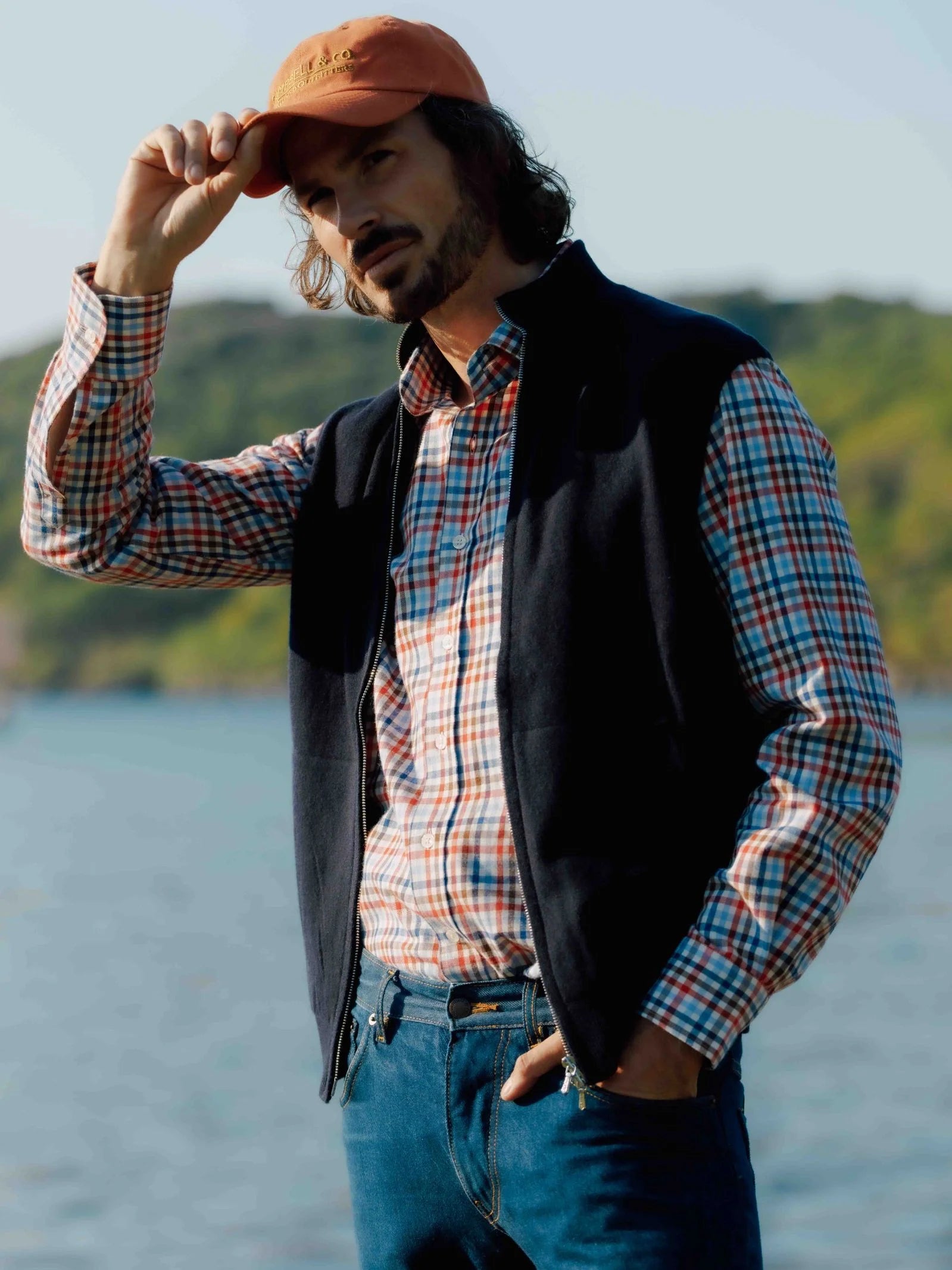 A man in a Campbell's of Beauly Windowpane Shirt, black vest, jeans, and an orange cap stands by the water outdoors, tilting his cap with one hand as he looks into the distance.