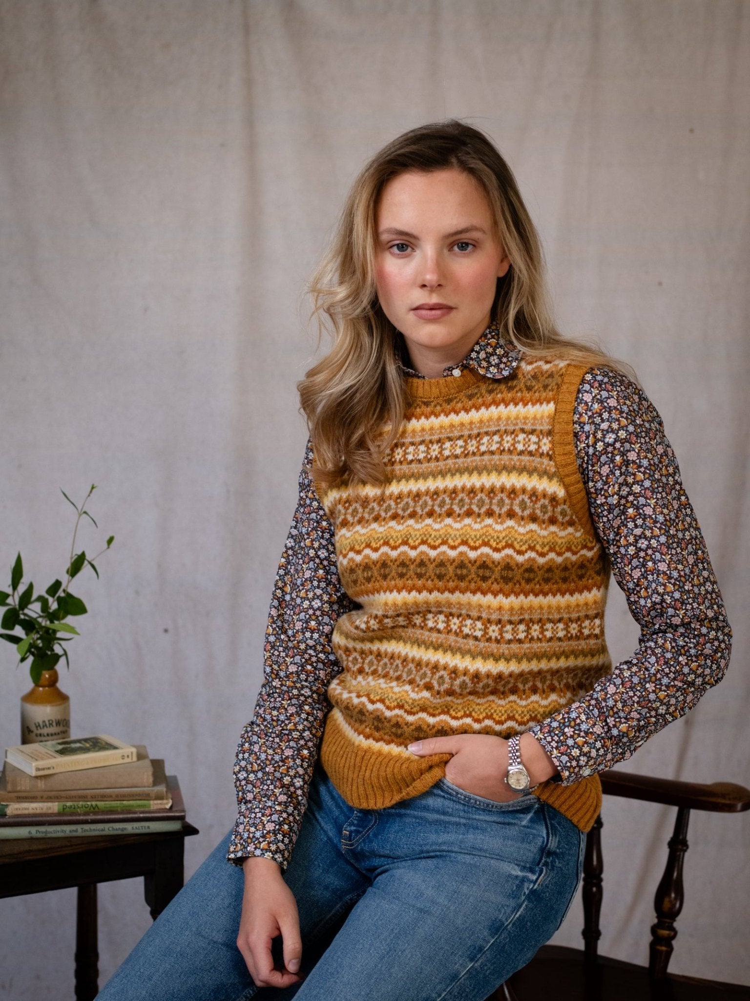 A woman with wavy blonde hair sits on a wooden chair, wearing the Campbell's of Beauly Fairisle Crew Tank over a floral shirt and blue jeans, her arm resting on a table with books and plants against a neutral fabric backdrop.